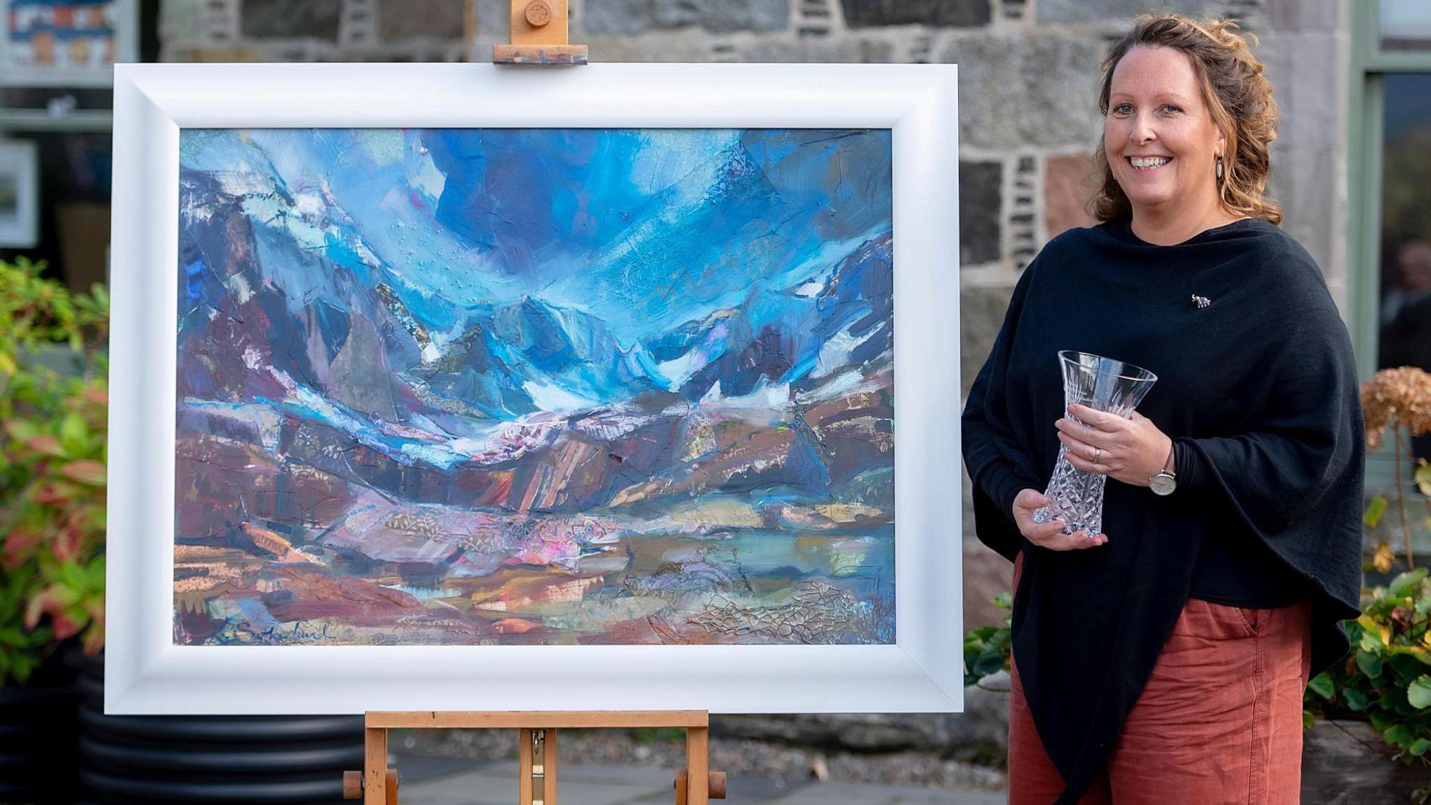 Katherine Sutherland is standing outdoors next to her painting of a mountain landscape. Katherine has brown hair, which is tied back, and is wearing a black outfit over a red skirt. She is holding the glass trophy she won.