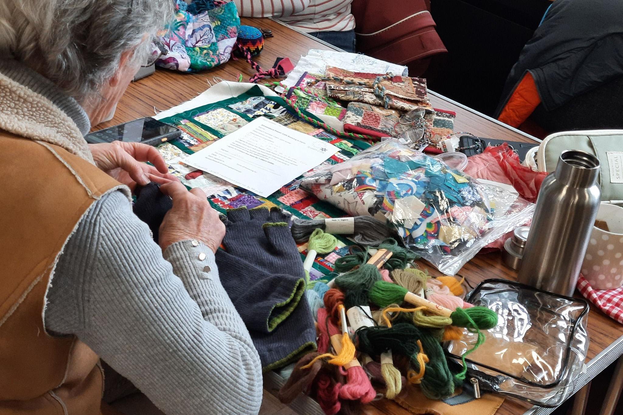 A table filled with different coloured threads and sewing equipment. A person's back can be seen sitting at the table, and the hands of volunteers on the other side.