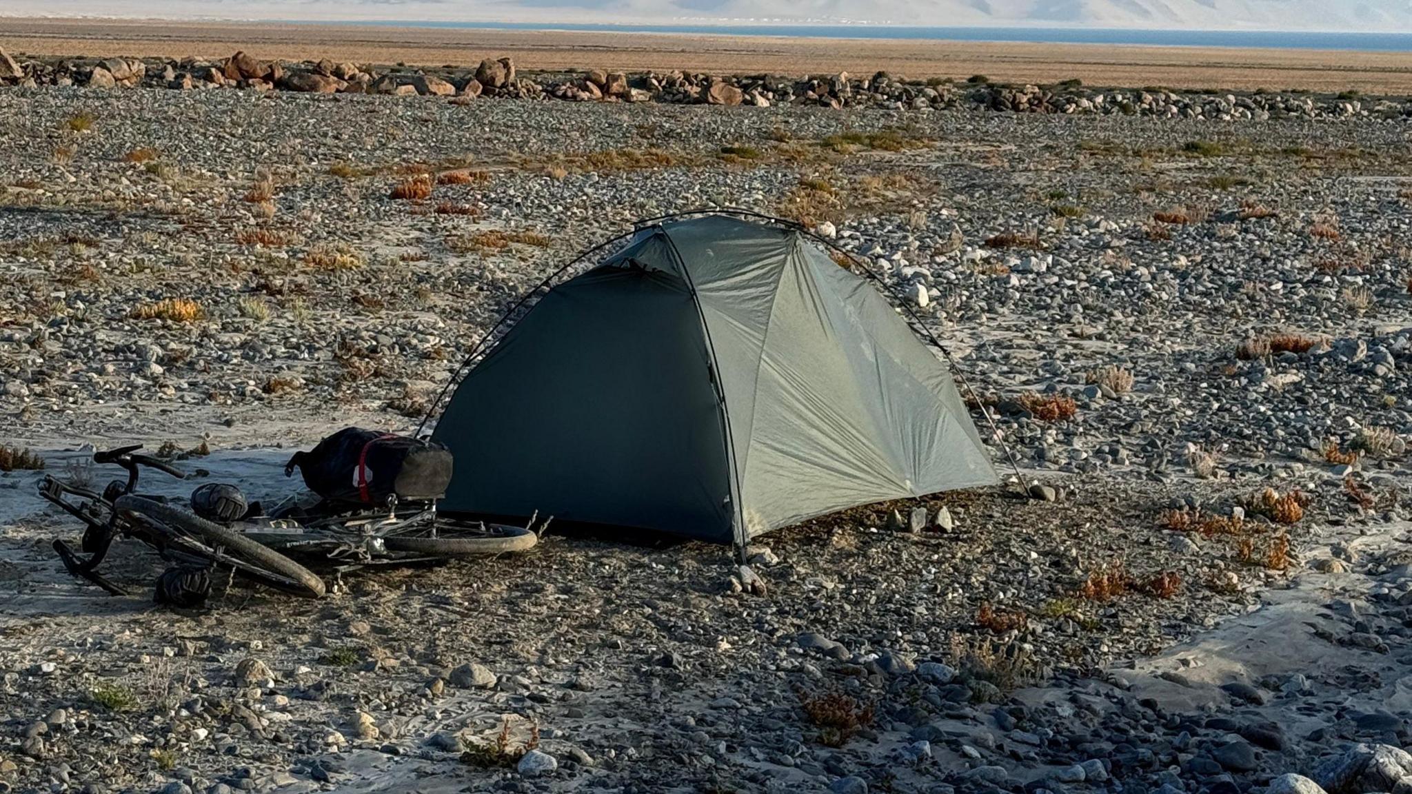 A dark green small tent with a bike on the floor next to it, standing on rough stony terrain, with dried grass in the background and then the sea.