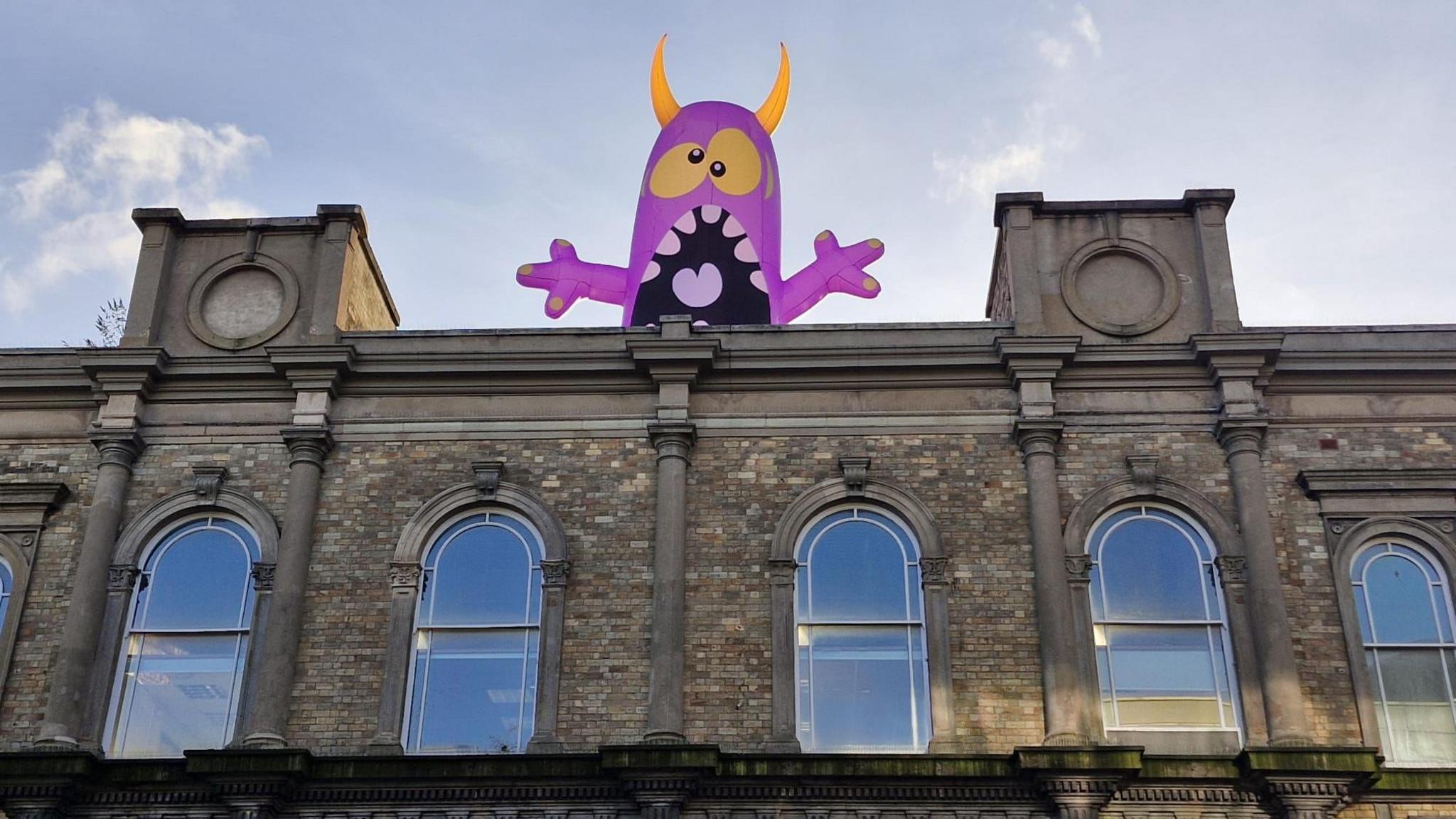 A purple inflatable monster with boggly eyes and big teeth looming over a building in Wolverhampton. There is a clear blue sky and the windows are grand and old fashioned.