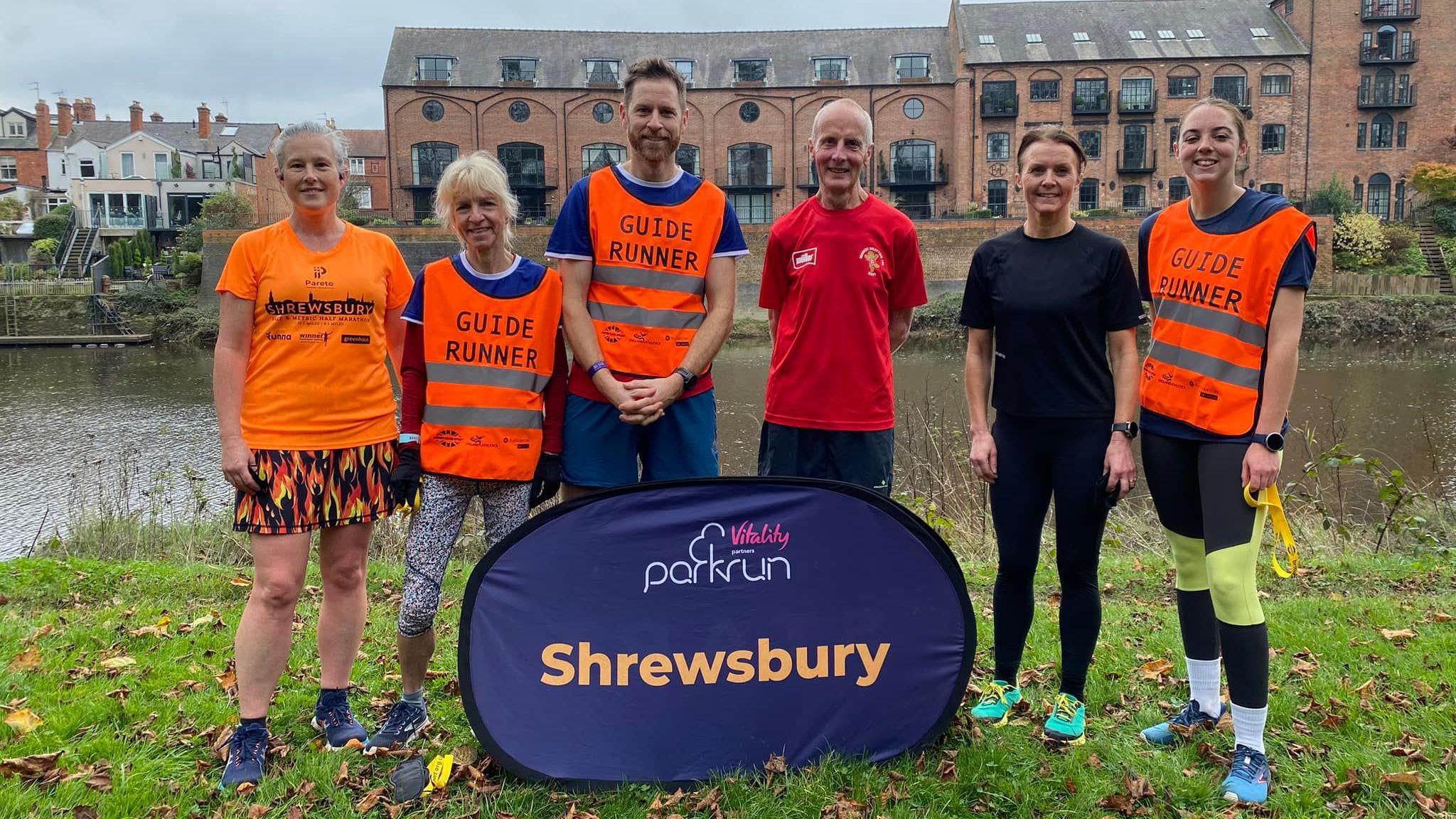 Sam, along with two women to his left and a man and two women to his right, are standing in front of the purple Shrewsbury parkrun banner, with the river Severn in the background. Sam and two of the women are wearing bright orange vests that say "guide runner".