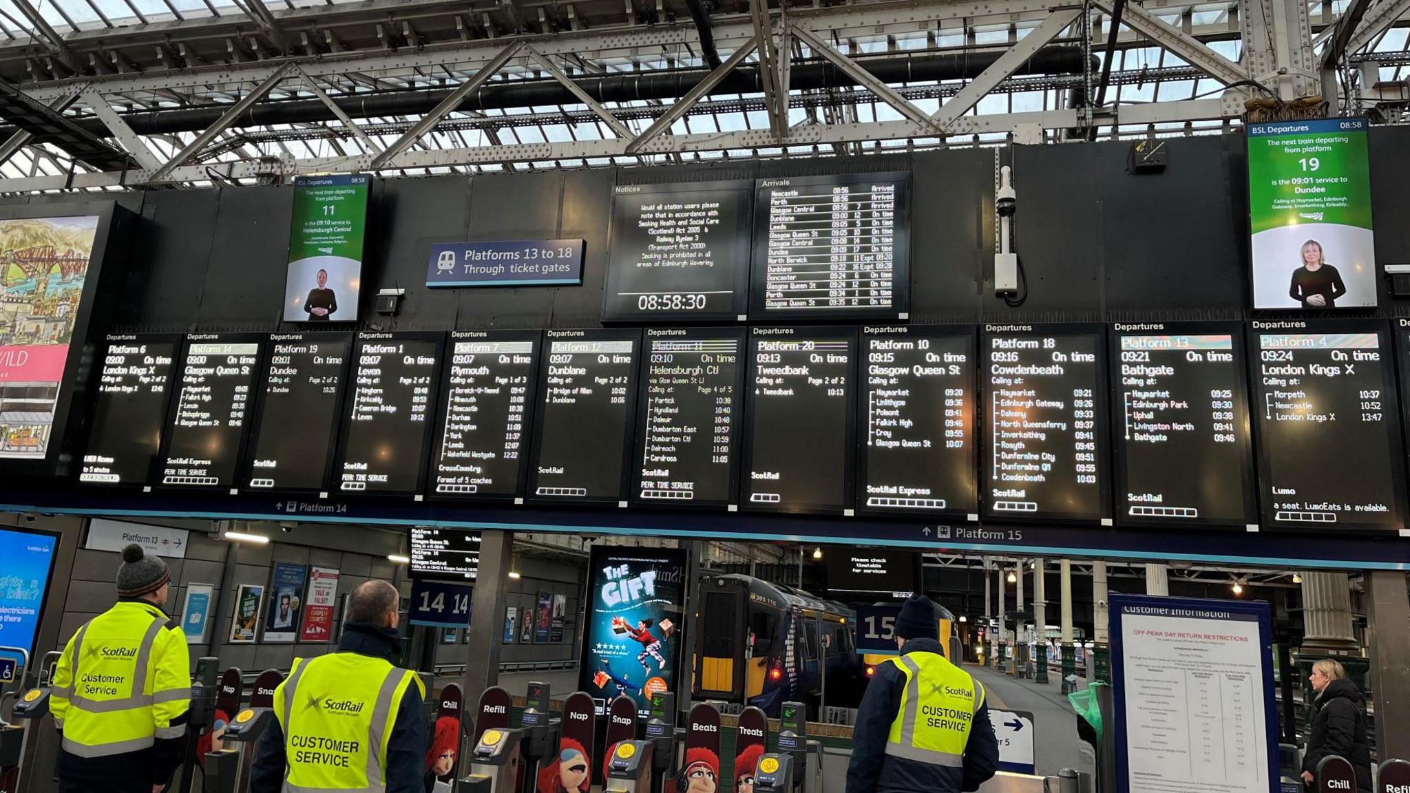 Image of three ScotRail operators in hi-vis jackets looking up at the departure board in Edinburgh Waverley, showing all services running as scheduled