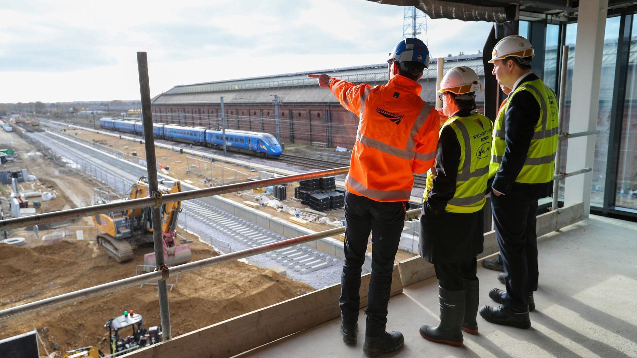 Luke Durston (Network Rail), councillor Steve Harker, Tees Valley Mayor Ben Houchen, and Steve Wilson (LNER) observe progress on the two new platforms. They are pictured from the back. Mr Durston is pointing at the building works. Tracks are being laid out and there is machinery. A blue  train is passing by. 