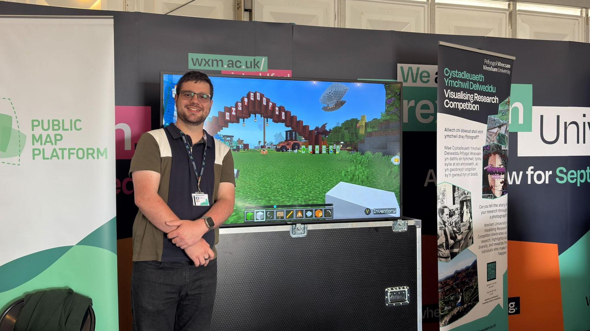 A brunette man wearing glasses and smiling towards the camera. He is wearing a patterned top and a lanyard, stood in front of a screen displaying the virtual Minecraft world of the Wrexham Eisteddfod.