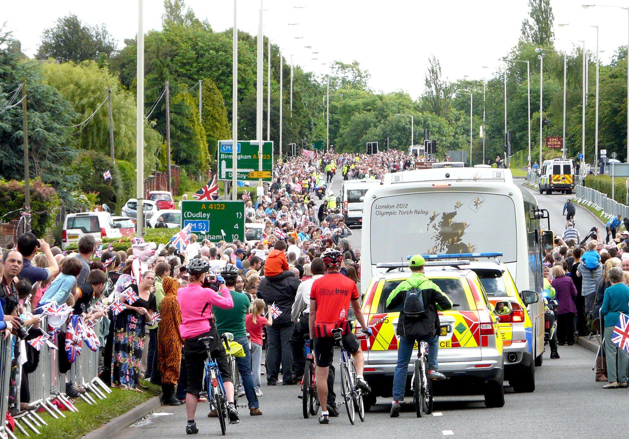 Crowds of union jack-waving onlookers watch as the torchbearer procession passes along the road. There are police vehicles and cycles behind a Torch Relay bus.