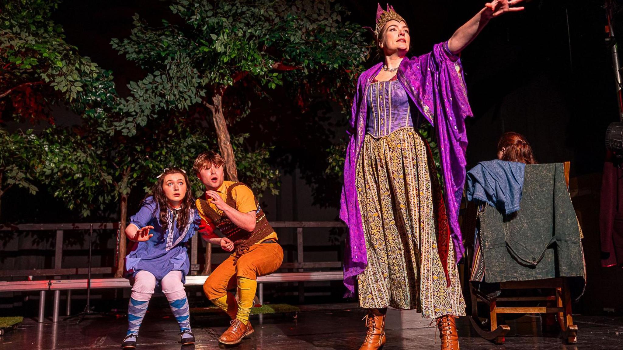 Three actors perform a scene on stage in East Belfast. In the foreground is a woman dressed as as witch in a paisley-patterned skirt, purple top and purple cloak and gold crown. Behind her two children cower - a girl with dark brown hair in a purple dress and purple tights and a boy with short brown hair in yellow socks, orange trousers, a yellow shirt and brown waistcoat