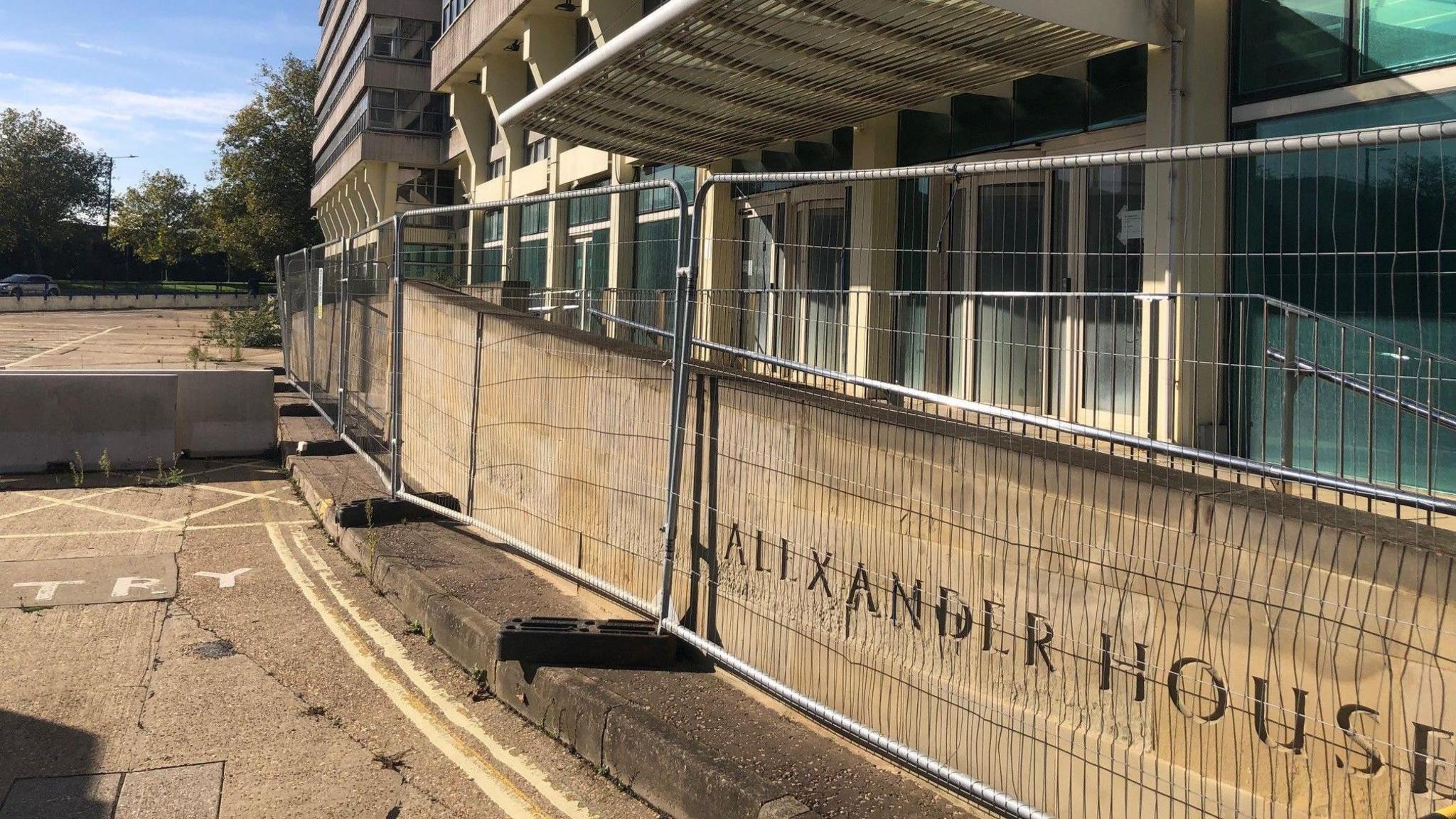 Another picture of Alexander House. It shows the ground floor, which is behind railings. A brick wall behind the railings has the name of the building engraved in it. The derelict car park can be seen in the background.