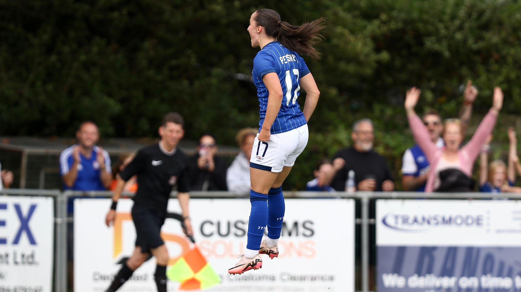 Sophie Peskett, jumping in celebration, wearing the blue Ipswich Town it, as fans raise their arms in the background. 