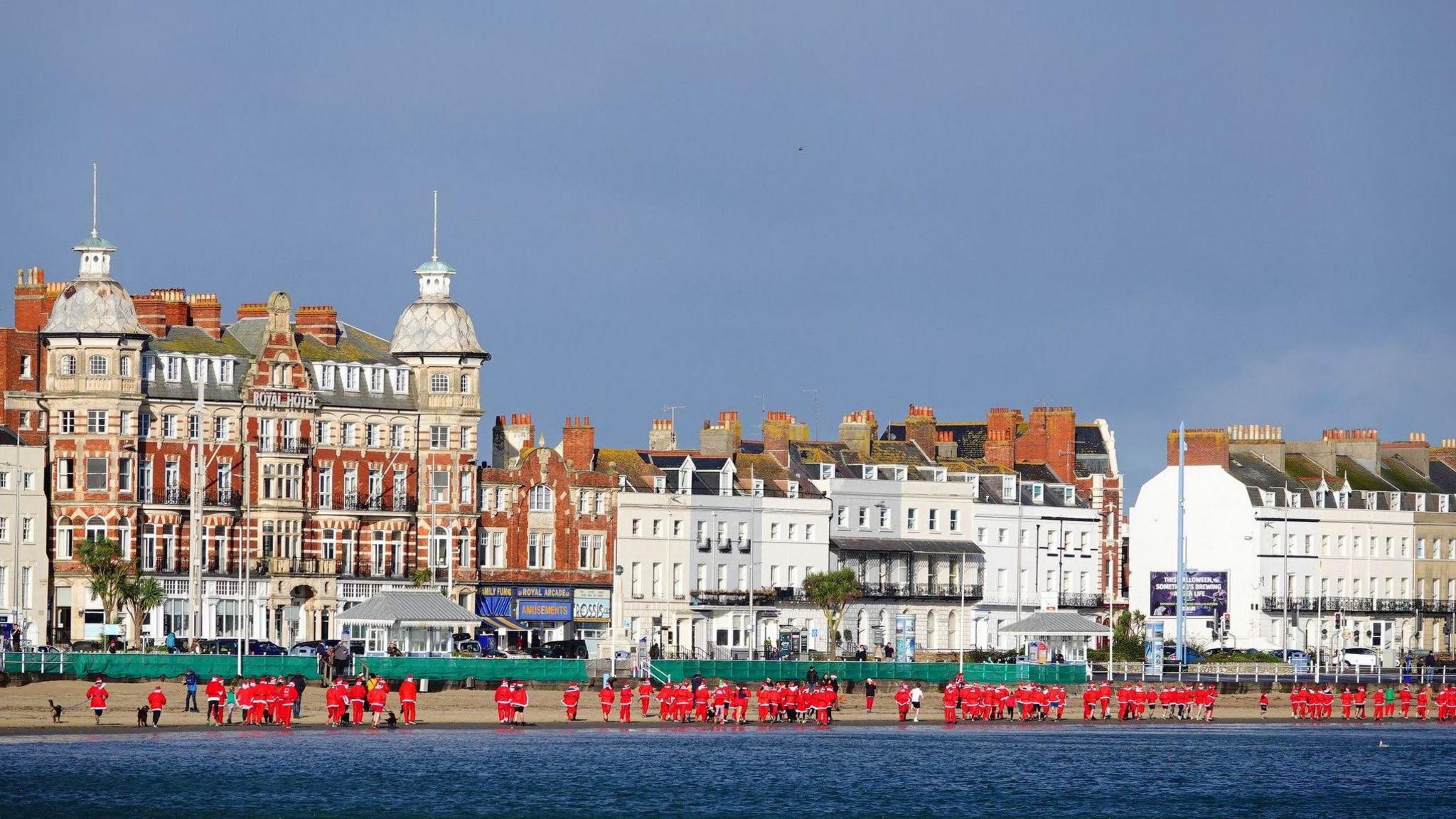 People dressed as santa on the beach from a distance in front of a row of buildings