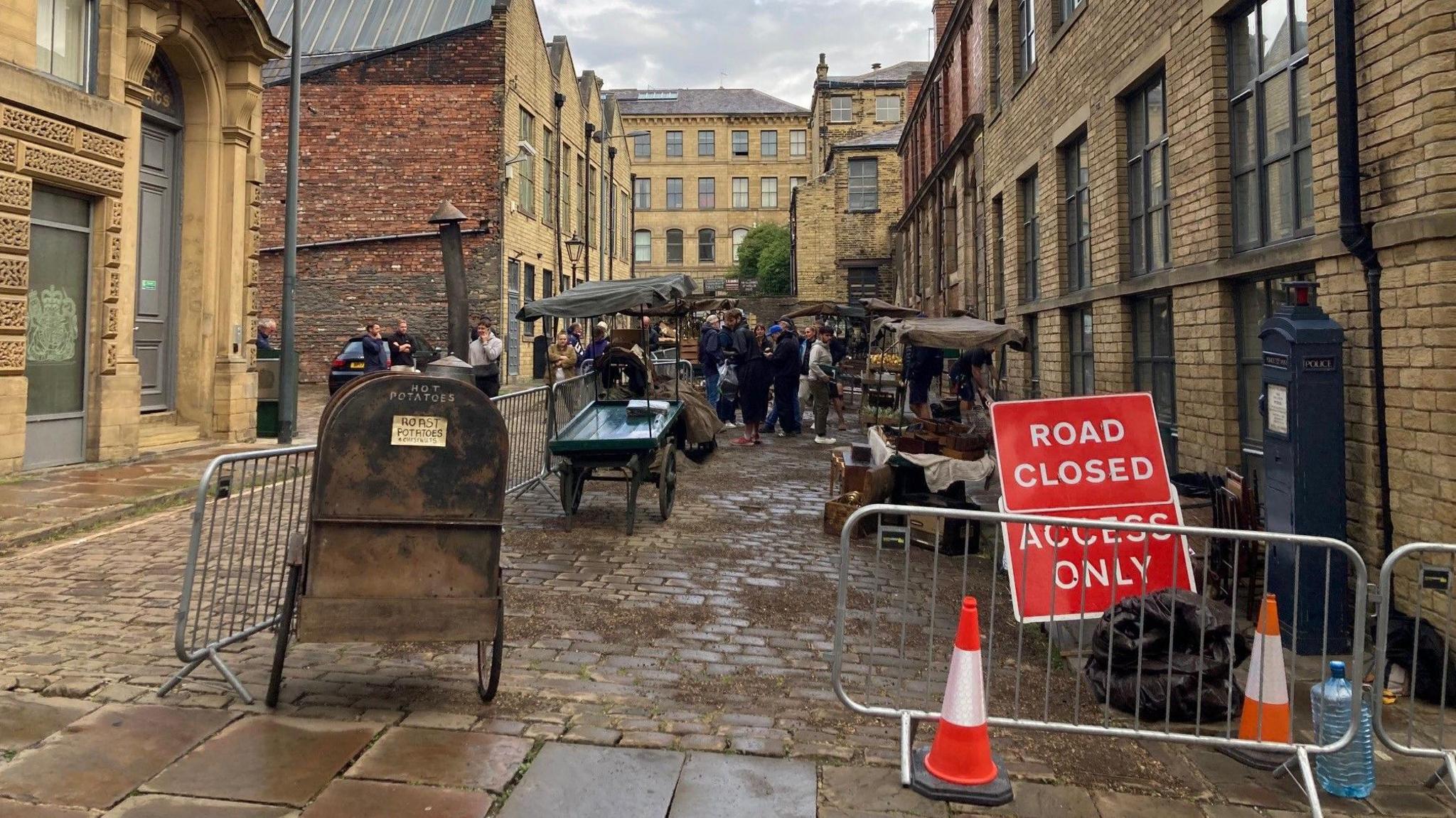 Metal railings around a street scene with old-fashioned wooden market stalls and a crowd of film crew members in the distance.   