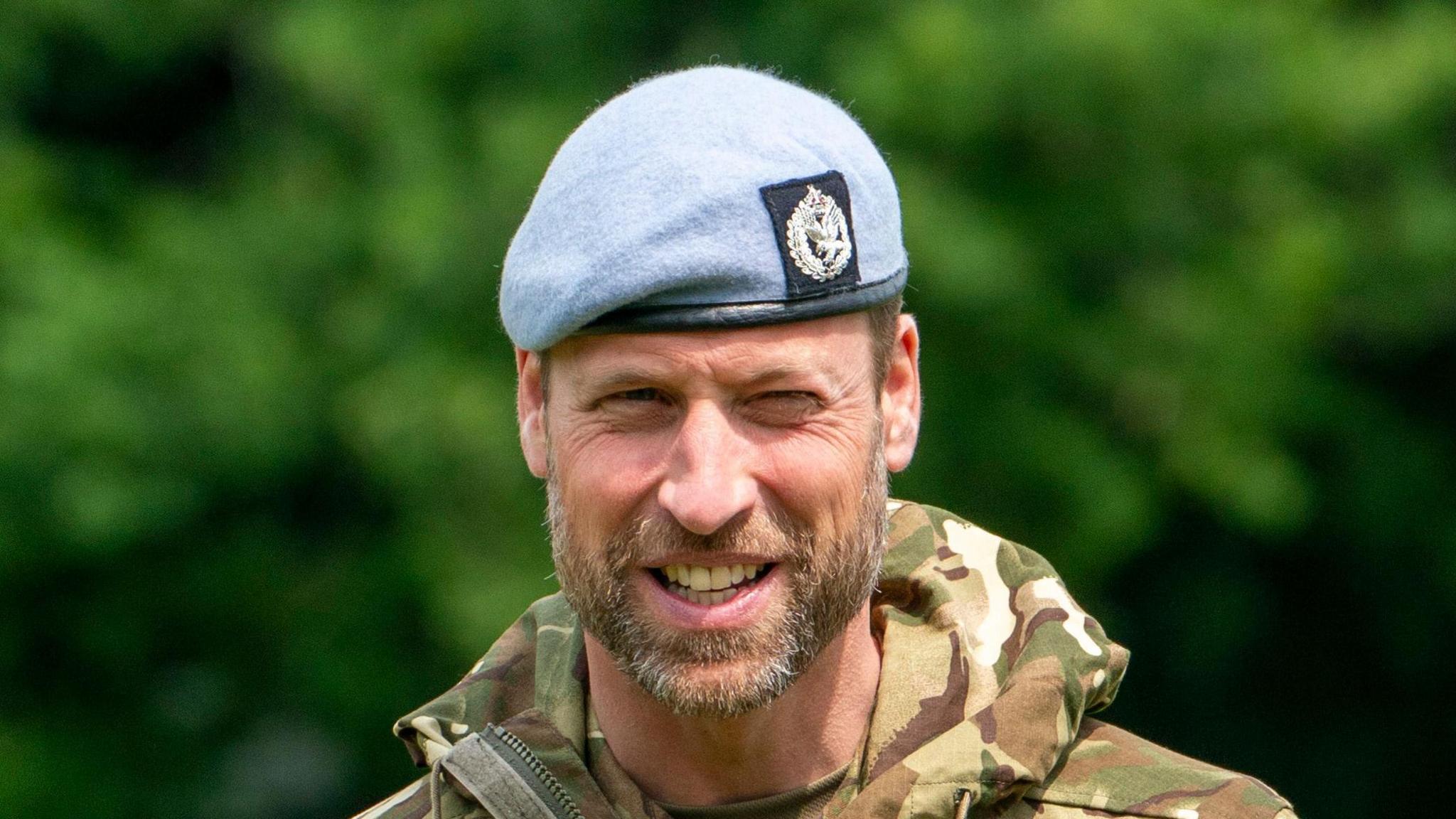 Prince William, stood outside, smiles and squints slightly. He wears his army uniform with a pale blue beret.