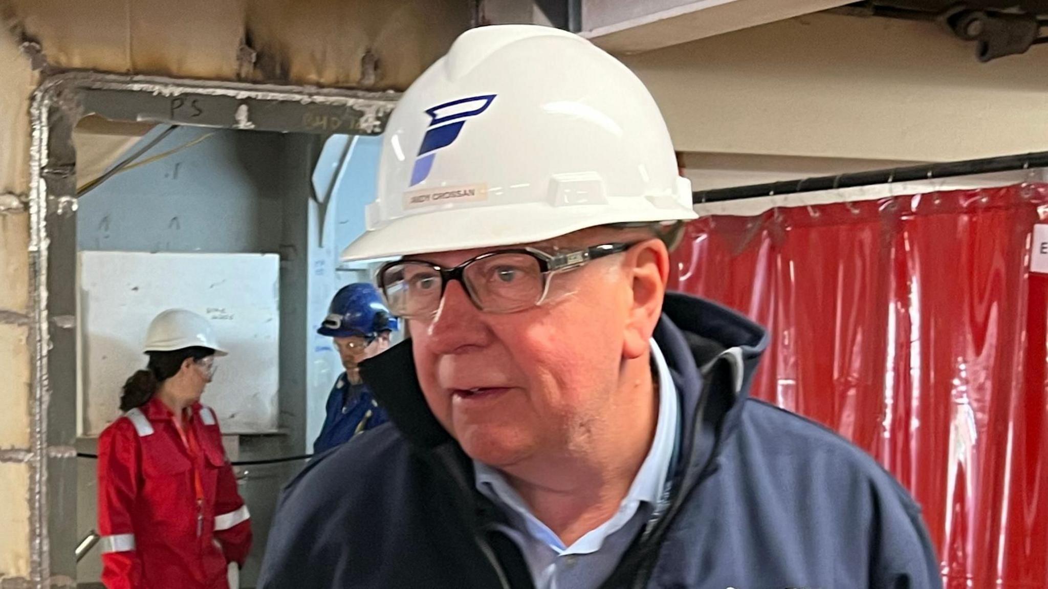 A bespectacled man wearing a dark jacket and a white helmet with the Ferguson Marine logo on the front