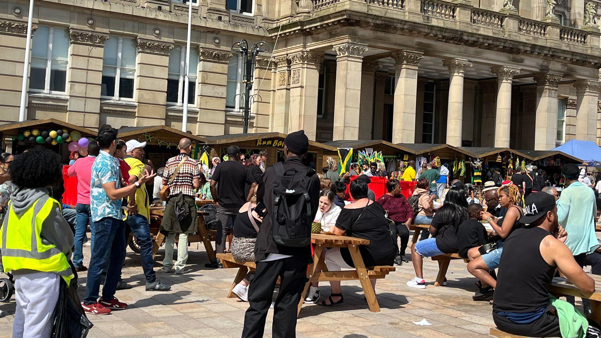 A crowd of people at the 0121 Jamaica festival, we see them thronged around park benches in front of several wooden stalls.