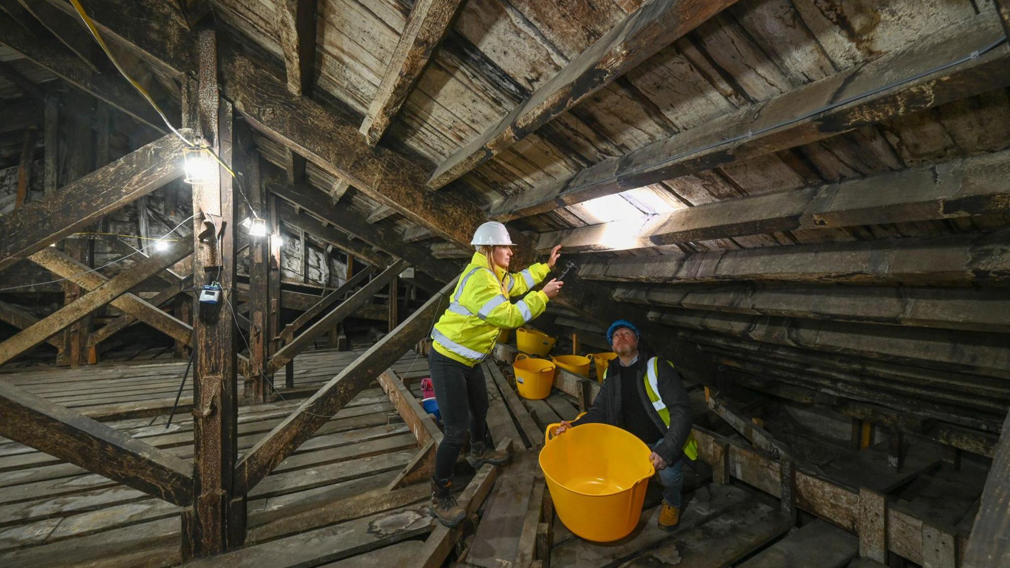 Two builders in yellow hi vis jackets work on the inside of Blenheim Palace's roof. They have several yellow buckets with them. One of them is shining a flashlight at the rood while standing up. The other one is sitting below and looking at the light.