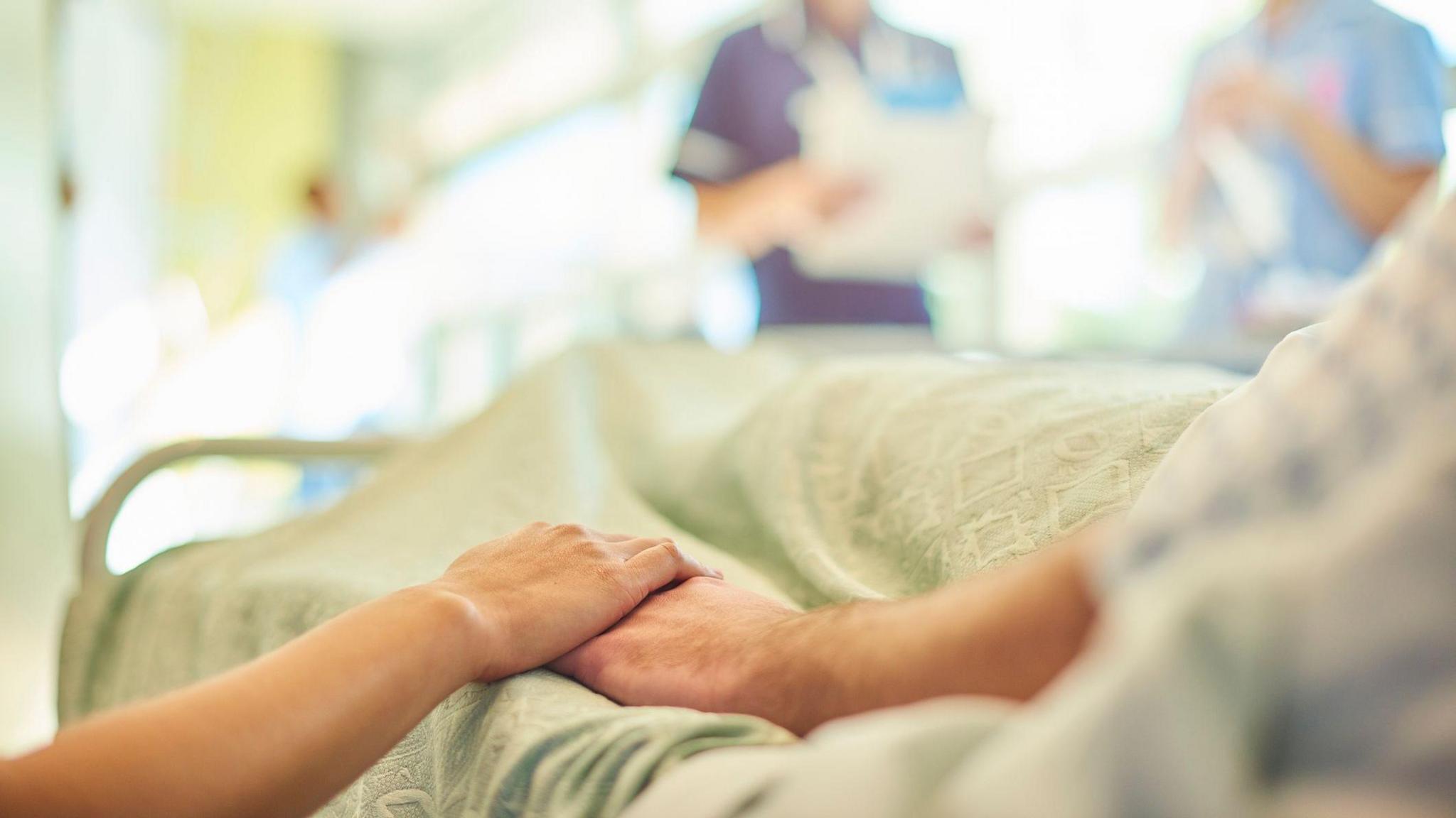 A hospital visitor's hand holds a patient's hand in bed of a hospital ward. In the blurred background a young nurse is chatting to the ward sister about the patient's care.