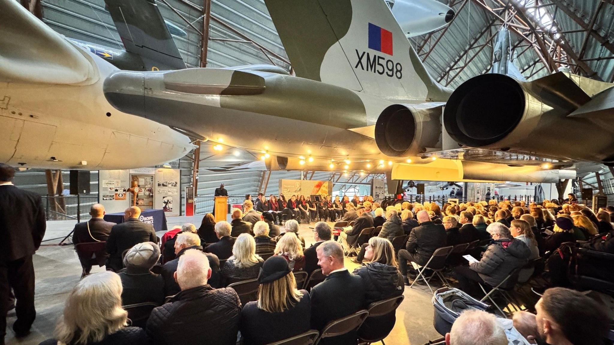 Rows of people in dark clothing sitting beneath the fuselage of a large military aircraft in a hangar