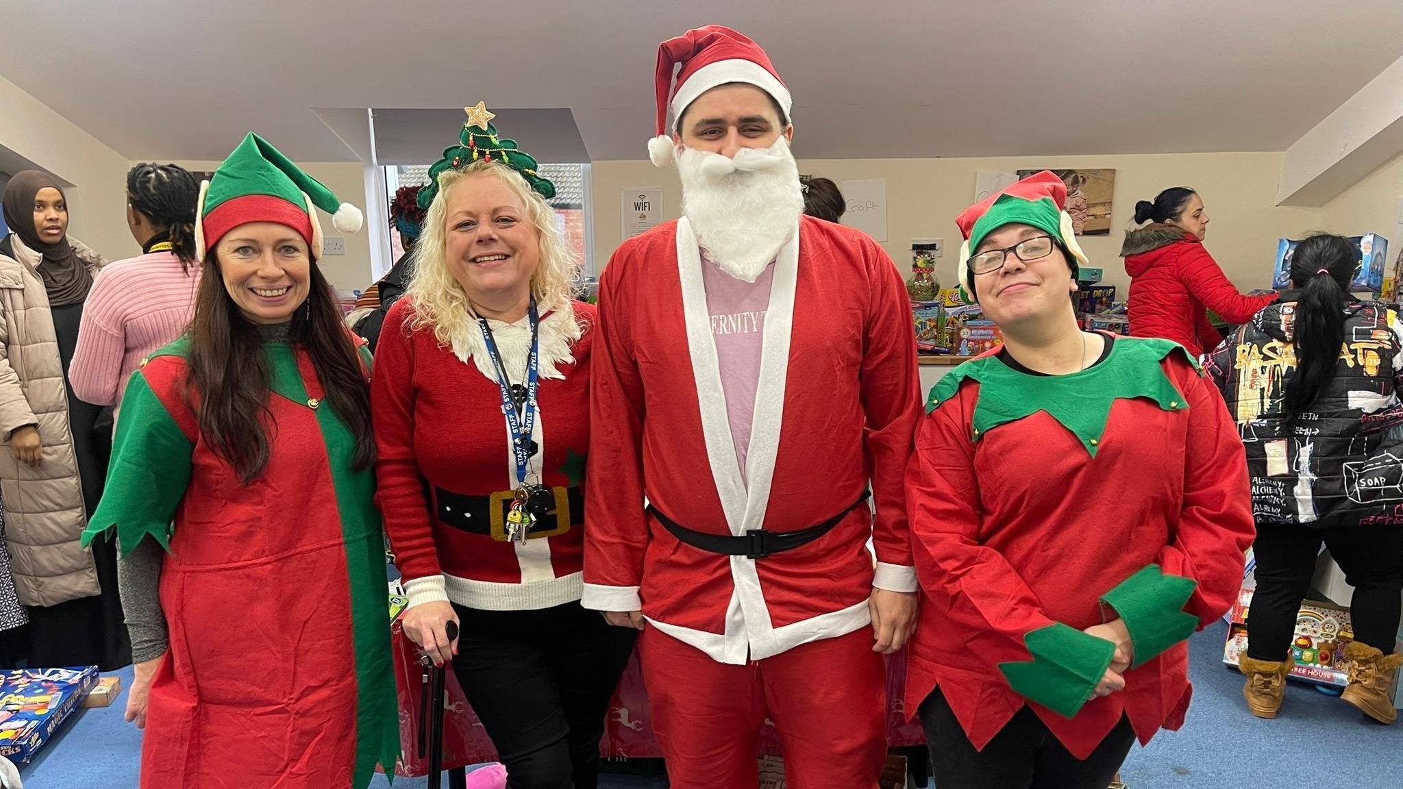 Four volunteers standing next to each other dressed in red and green Christmas apparel. Two people are dressed as elves while two others have donned a santa suit. 