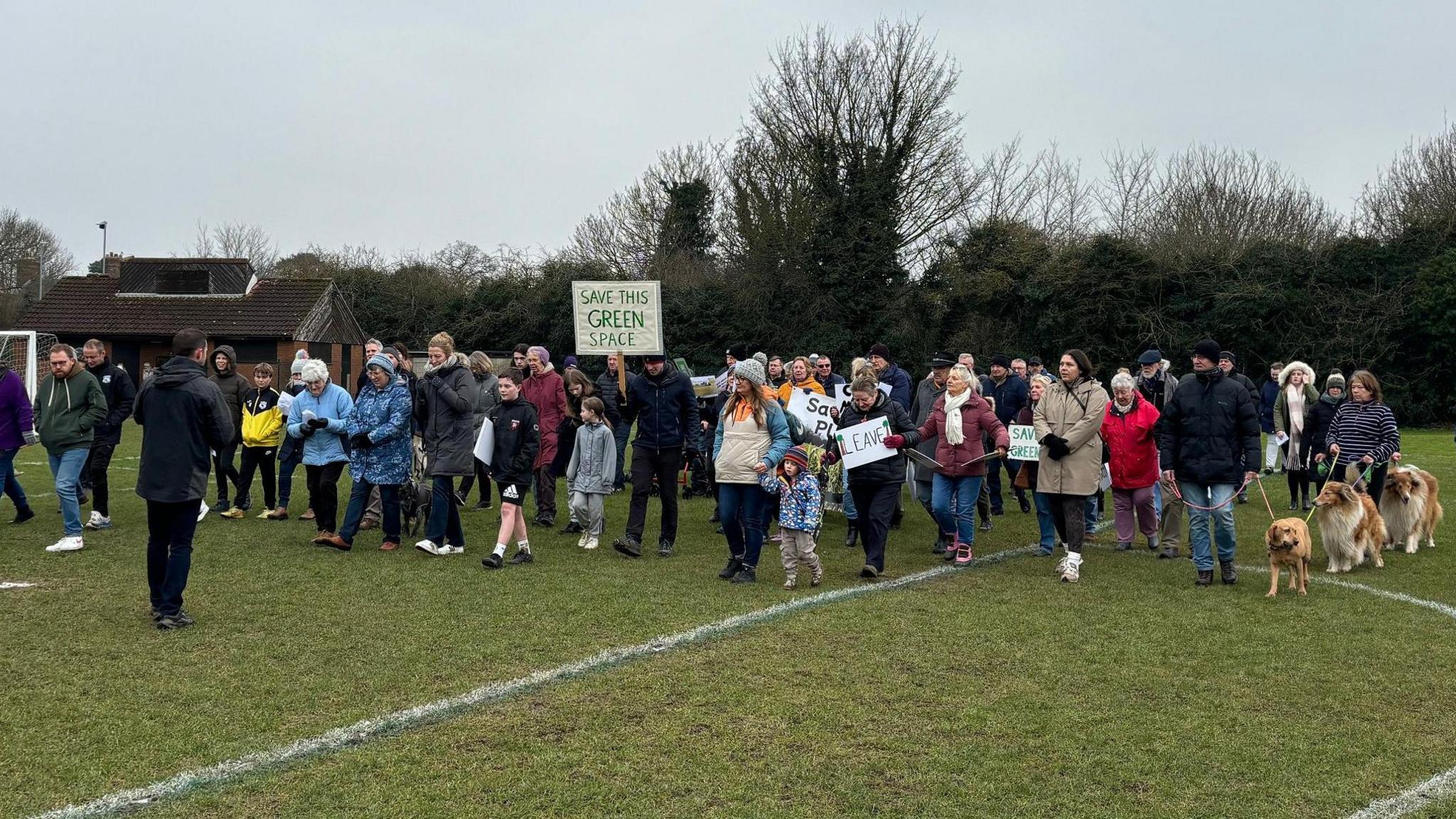 A group of about 50 people including children are walking together across a football field. Some are holding placards reading things like 'leave' or 'save this green space'. Some people have dogs on leads. 