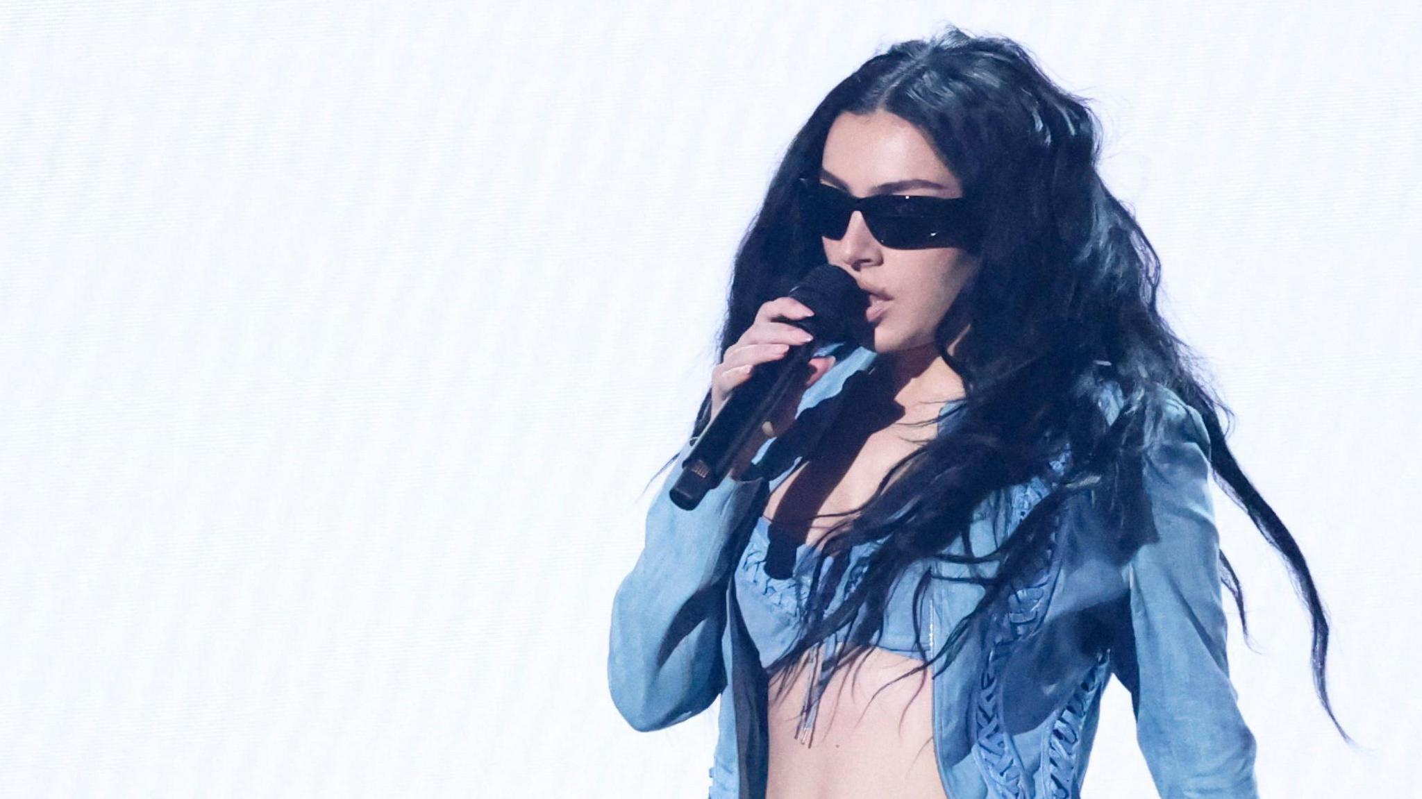 Charli XCX in a denim jacket and bra-top, sings into a microphone during the 2025 Grammy Awards