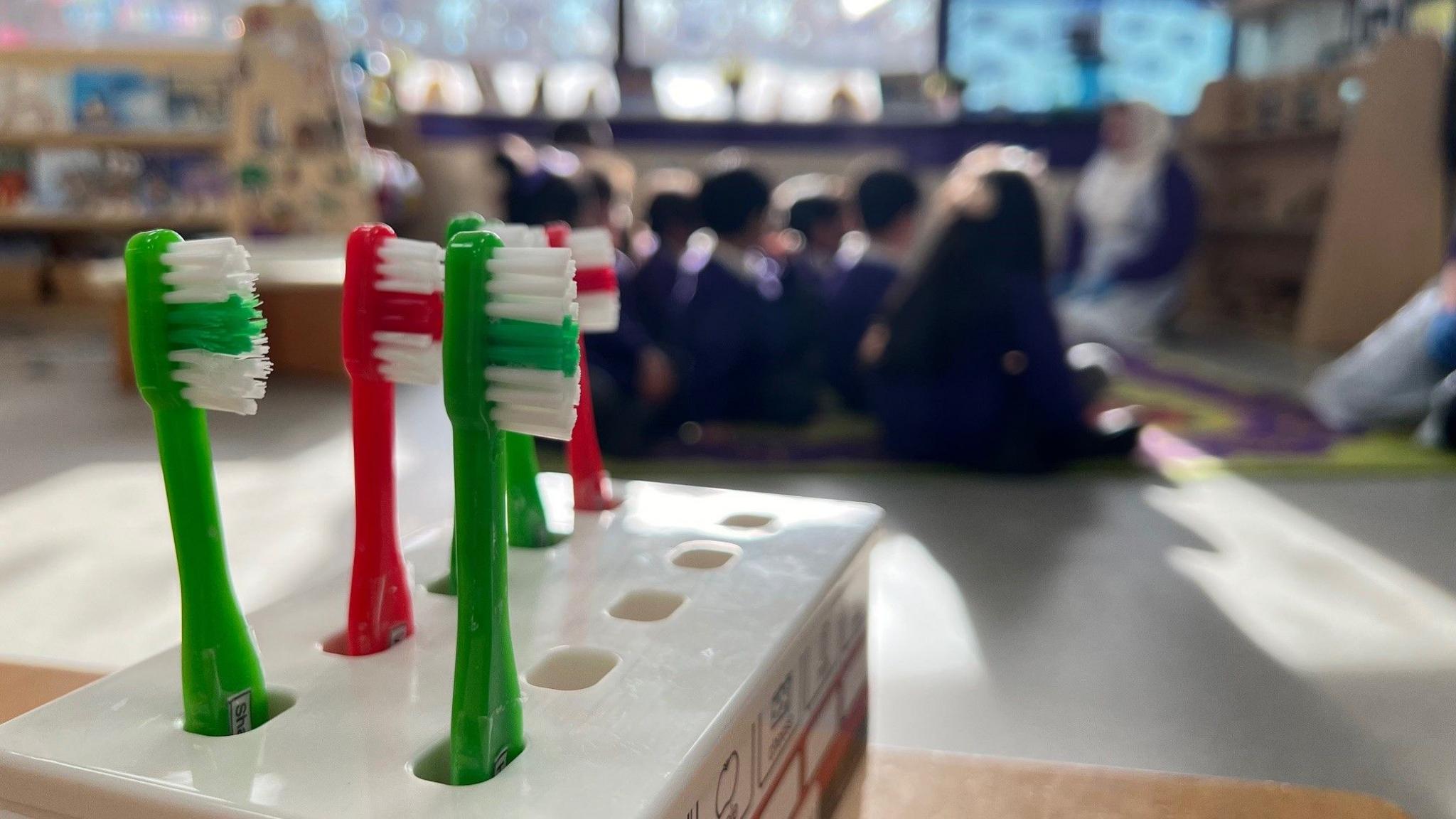 In the foreground there is a toothbrush holder.  It contains three green and two red toothbrushes. In the background, slightly out of focus, are a group of school children who are sitting on a rug and looking towards their teacher.