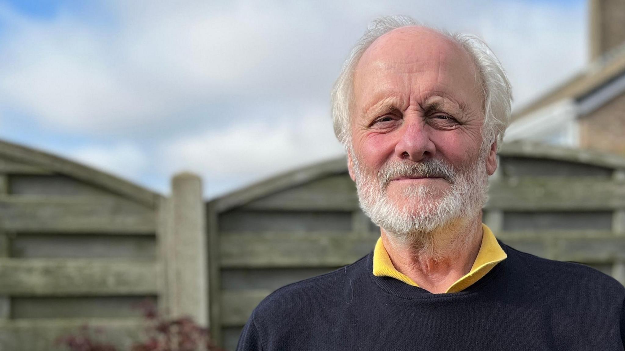 Chris Feast stands in his back garden in front of a grey wooden fence and a cloudy blue sky. He has white hair and a white beard and is wearing a black jumper over a yellow shirt.