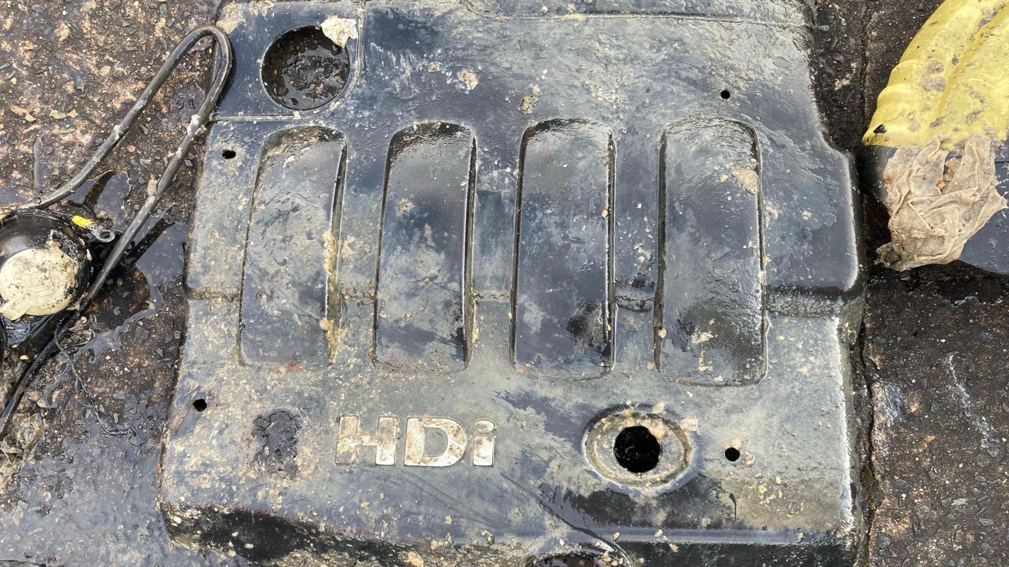 A vehicle engine cover. It is dirty, having been recovered from a sewer. It is black. It contains the letters "HDi".