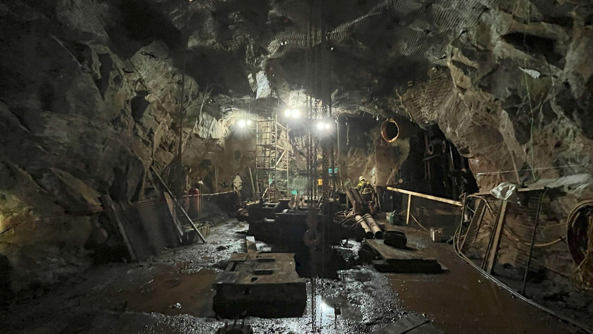 A dark cavern below ground all around is heavy equipment, chains and scaffolding. Floodlights illuminate the scene. 