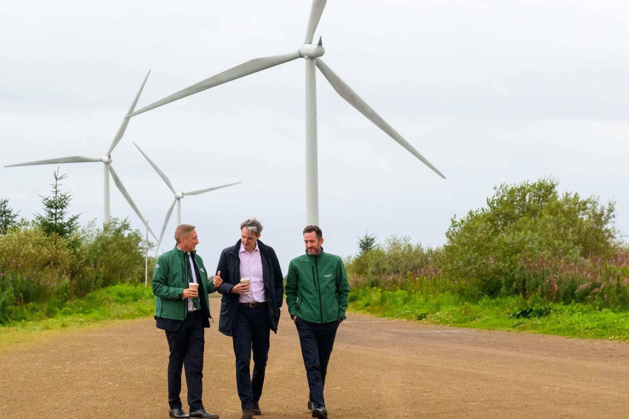 Paul Thwaite walks holding a coffee in between two men wearing green jackets. Behind them, you can see wind turbine and a wide grit path with some trees.