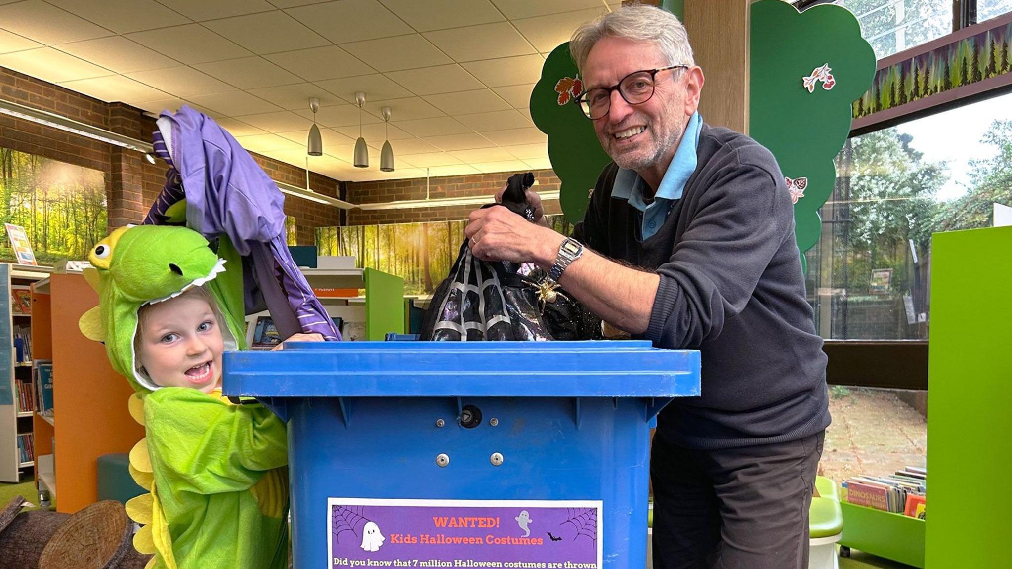 A smiling young girl dressed up as a green dinosaur is donating her old Halloween costumes at a local library. She is indoors, standing next to a blue wheelie bin, with a sign on that says 'Wanted! Kids Halloween Costumes'. On the opposite side of the bin is the library supervisor - he is an older man wearing glasses, dark trousers and a dark jumper over a blue shirt. He is also putting a donation in the bin and smiling at the camera. 