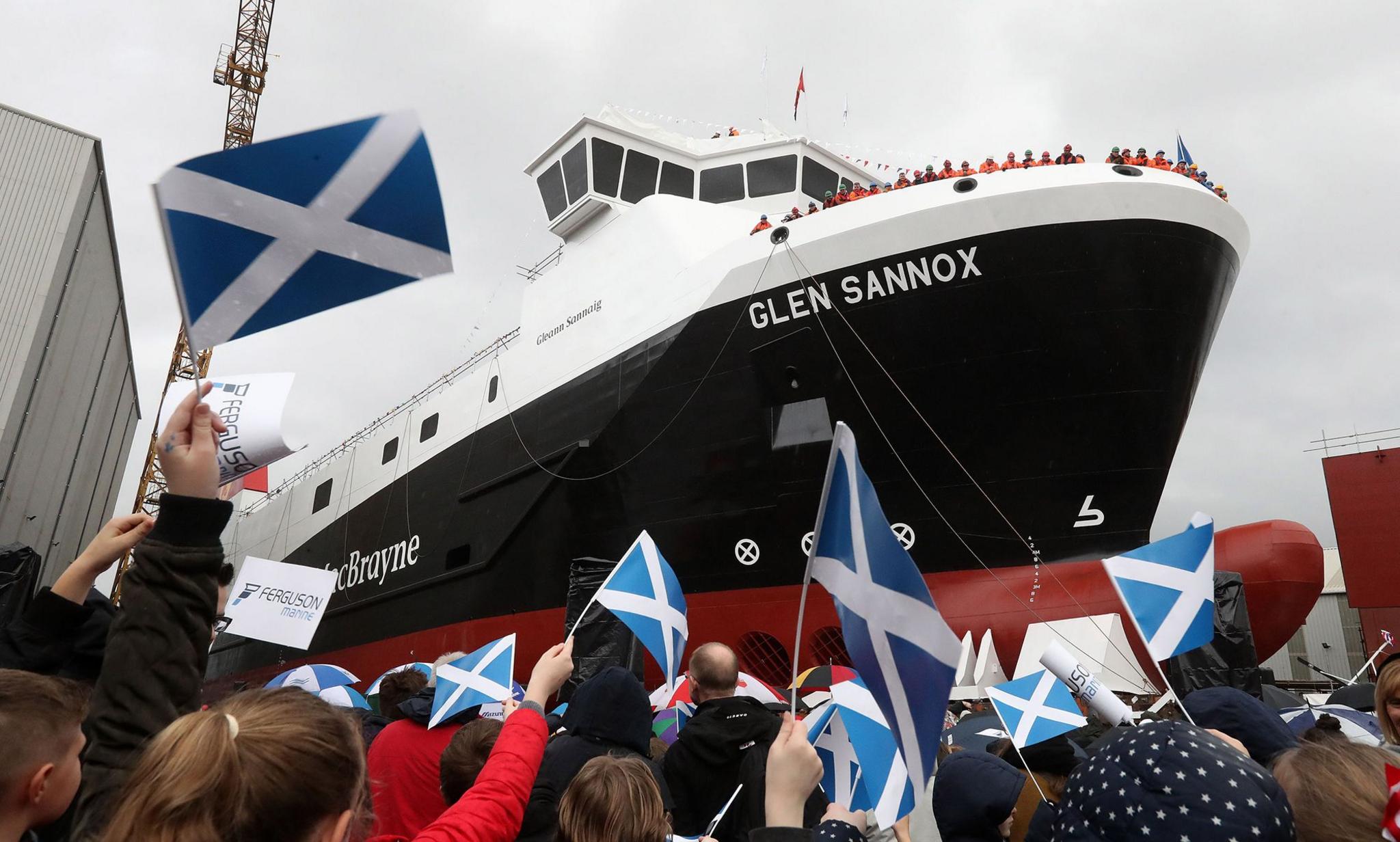 People wave saltires while watching a large red, black and white ship move down the slipway.