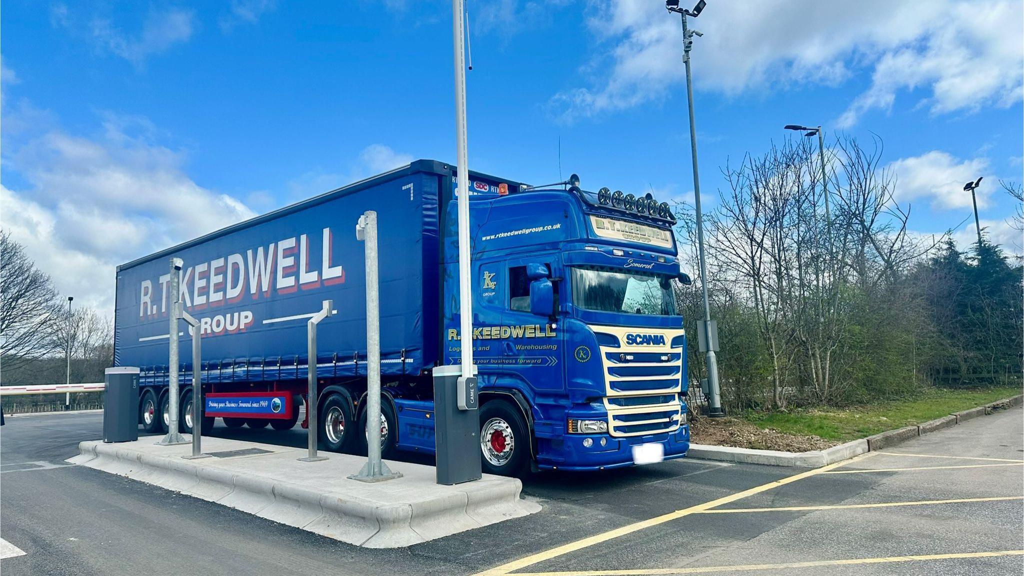 A large blue lorry coming through a barrier into a new parking area, under a blue sky.