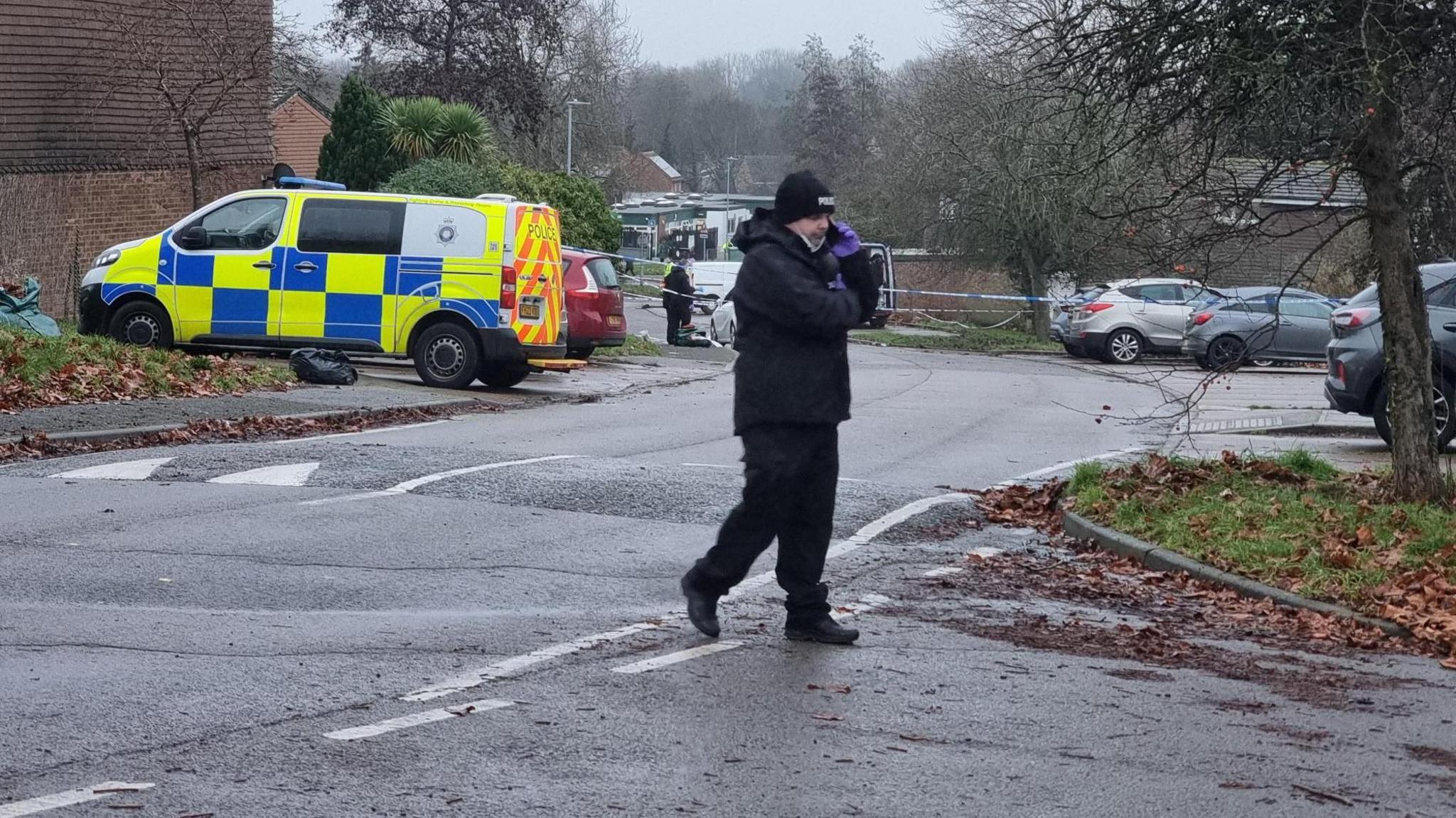 A police officer in black coat, hat and trousers walks across a road on the phone. A police van and police tape is across the road.