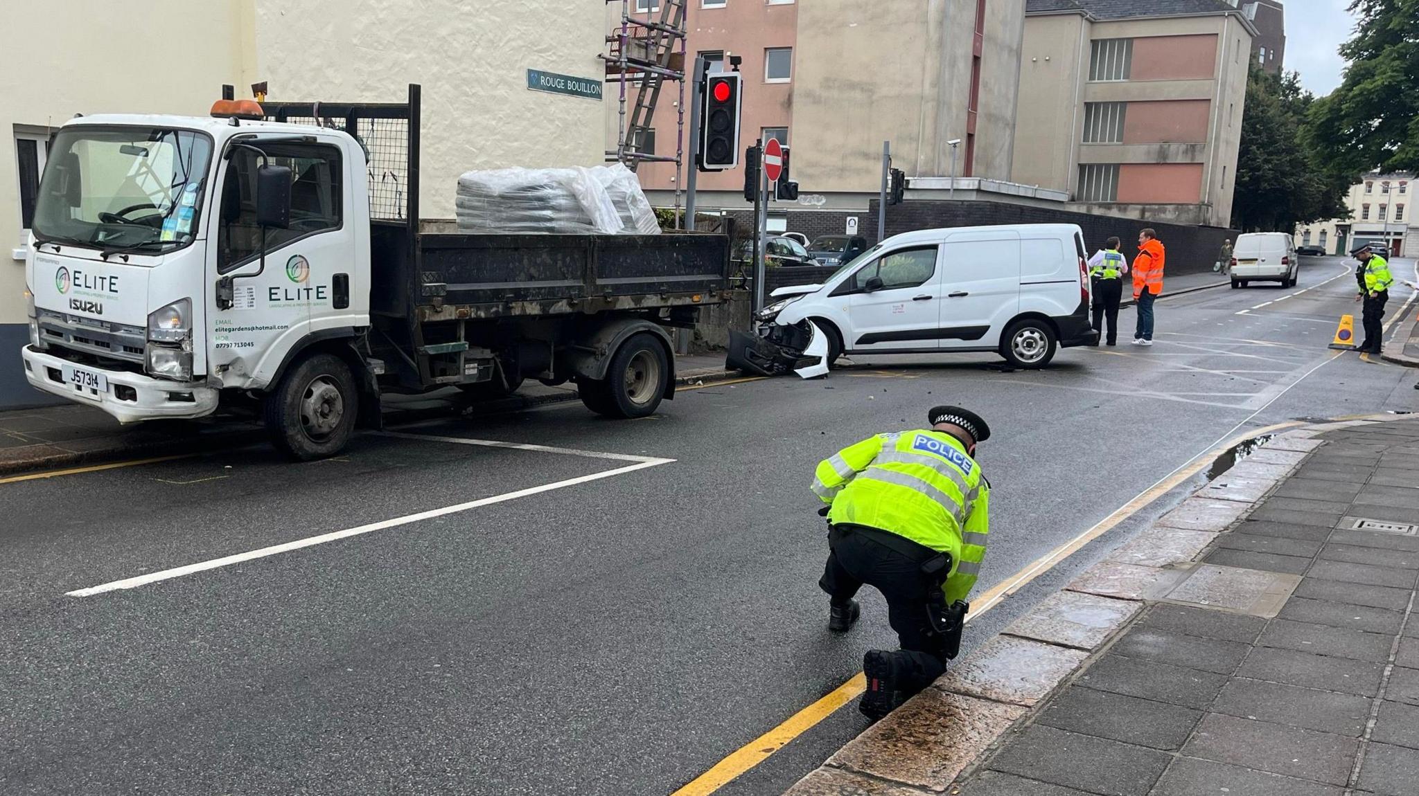 A small white van and a larger white truck. Both have damage to the front left of their chassis. The smaller van has hit a stop sign. A policeman kneels on the road.