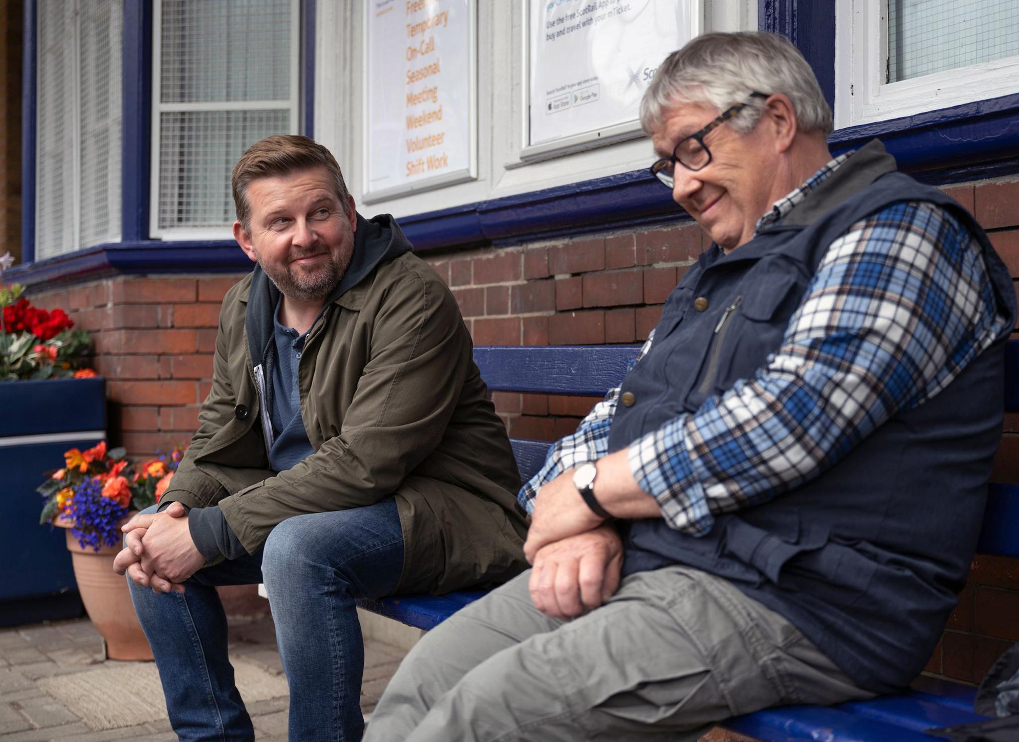 A bearded younger man sits on a station bench beside an older man dressed in a check shirt