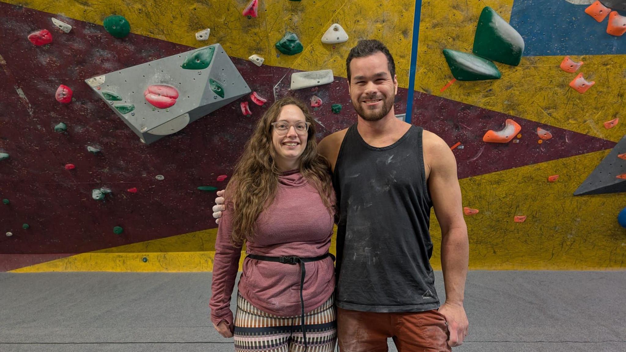 A woman and a man stand in front of a multicolour climbing wall. The man's arm is round the woman. They are both smiling at the camera and the man has white chalk on his clothes