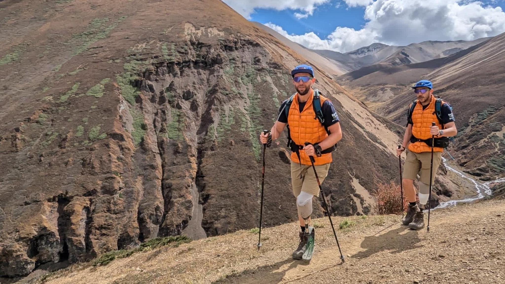 Rich Potter and Rich Davies wearing hats, sunglasses, orange and black short-sleeved tops, light brown shorts and black boots walking through a hilly area. Each has an artificial left leg and both are using trekking poles.