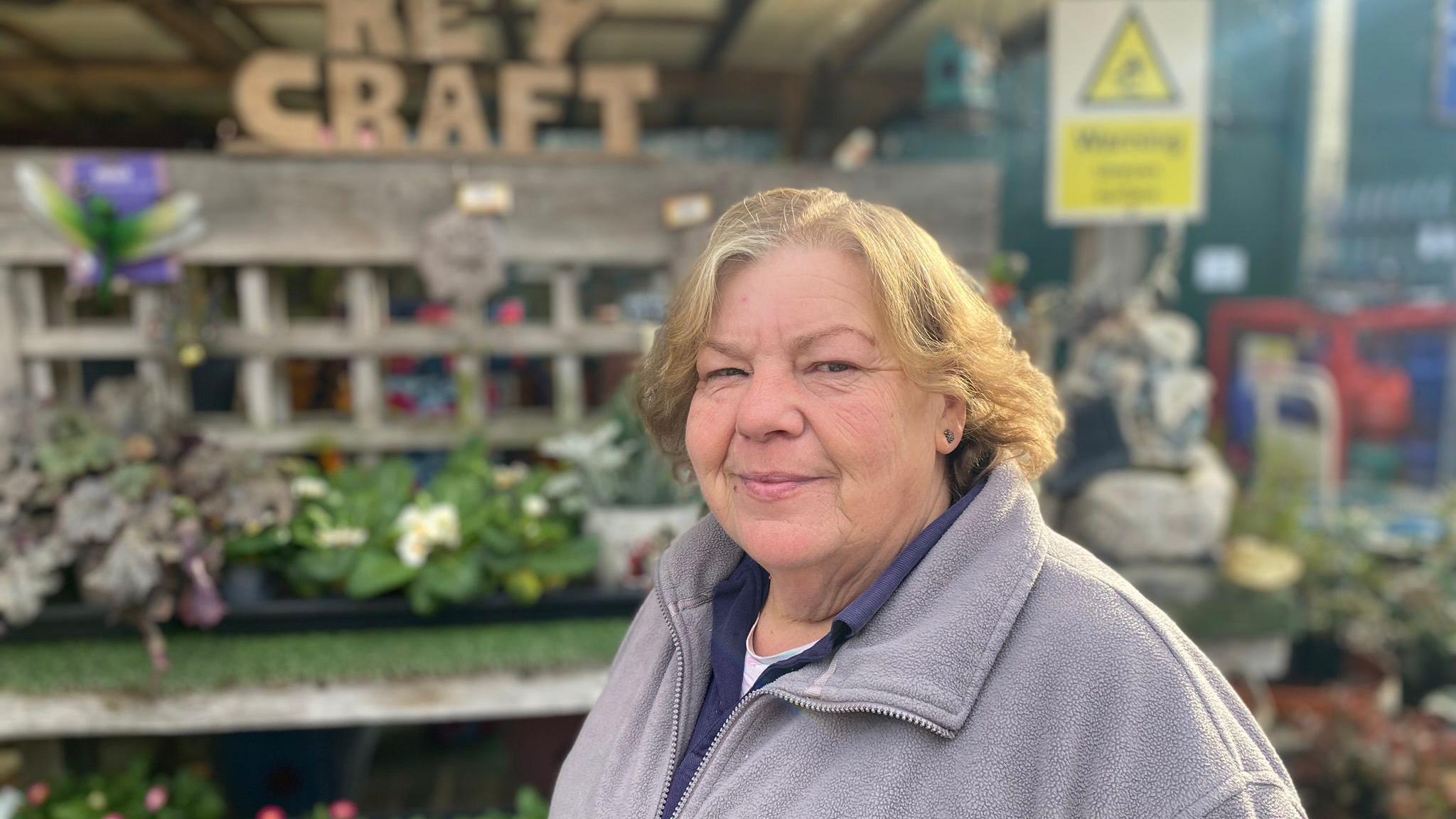 Jackie Wells, wearing a fleece, looking at the camera while stood in front of the Key Craft garden centre, which has plants outside