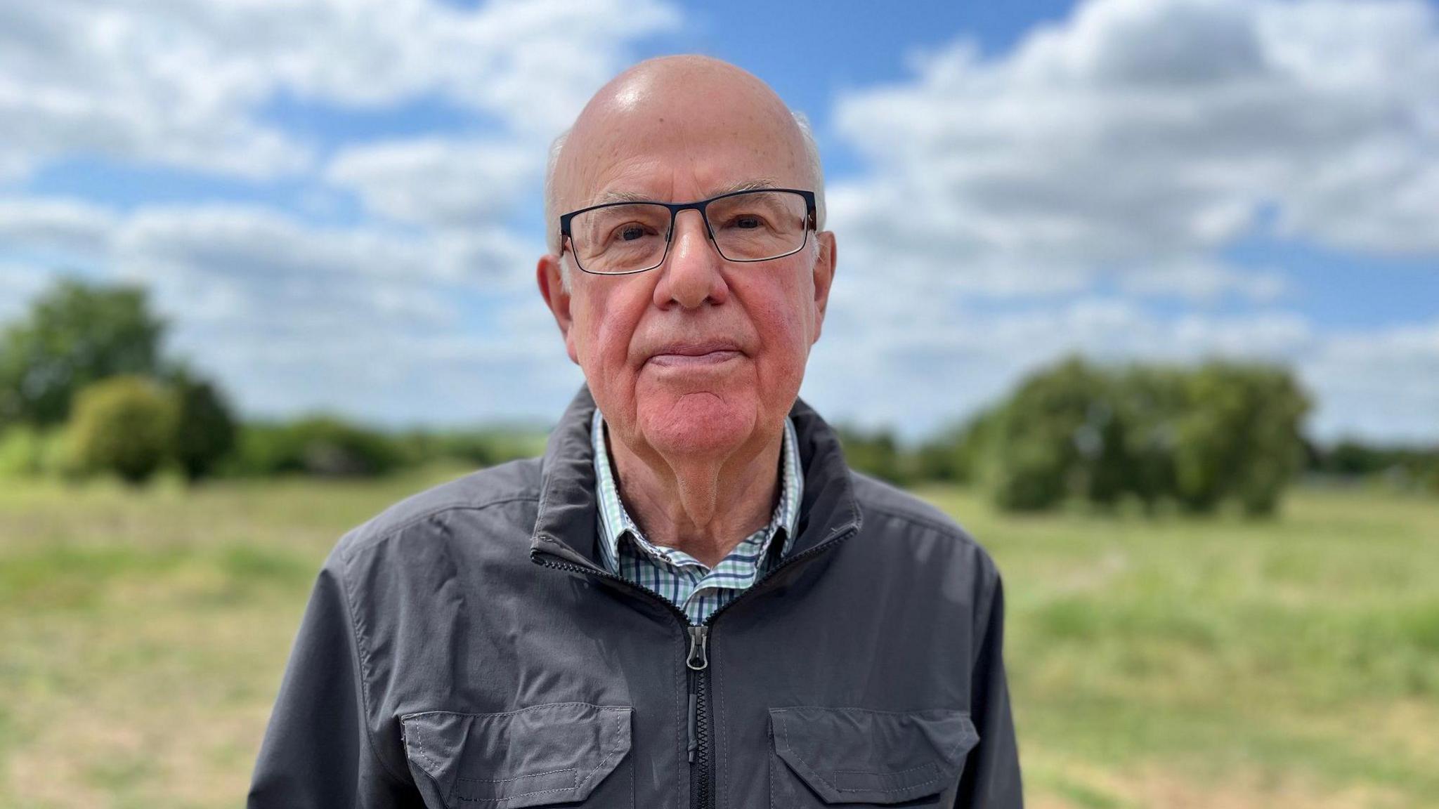 Robin Clegg looks into the camera wearing glasses and a blue jacket. He stands in front of the site, green with trees.