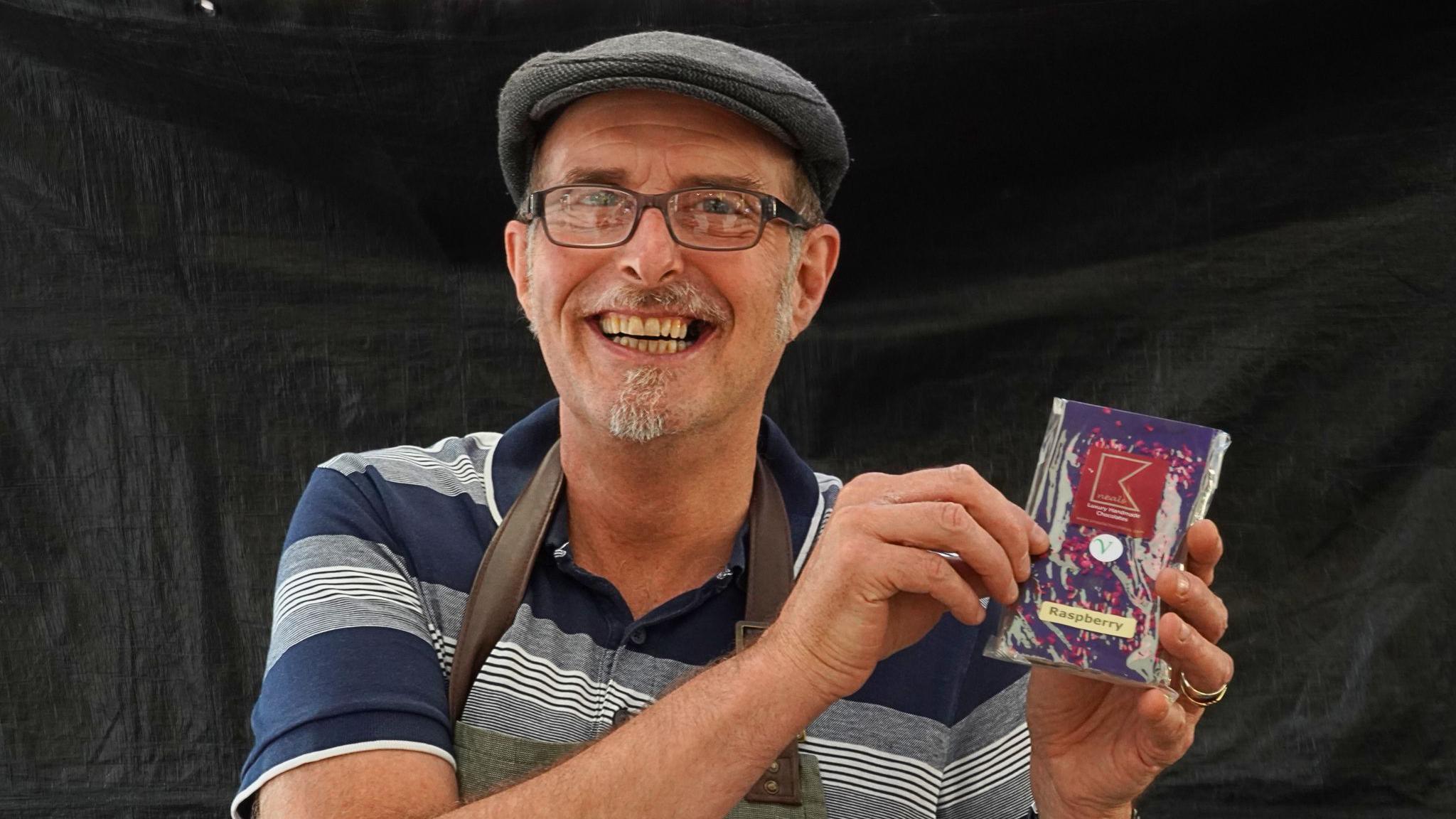 A man in a flat cap, who is wearing a navy blue striped polo top is holding a small box of chocolates and smiling at the camera. 