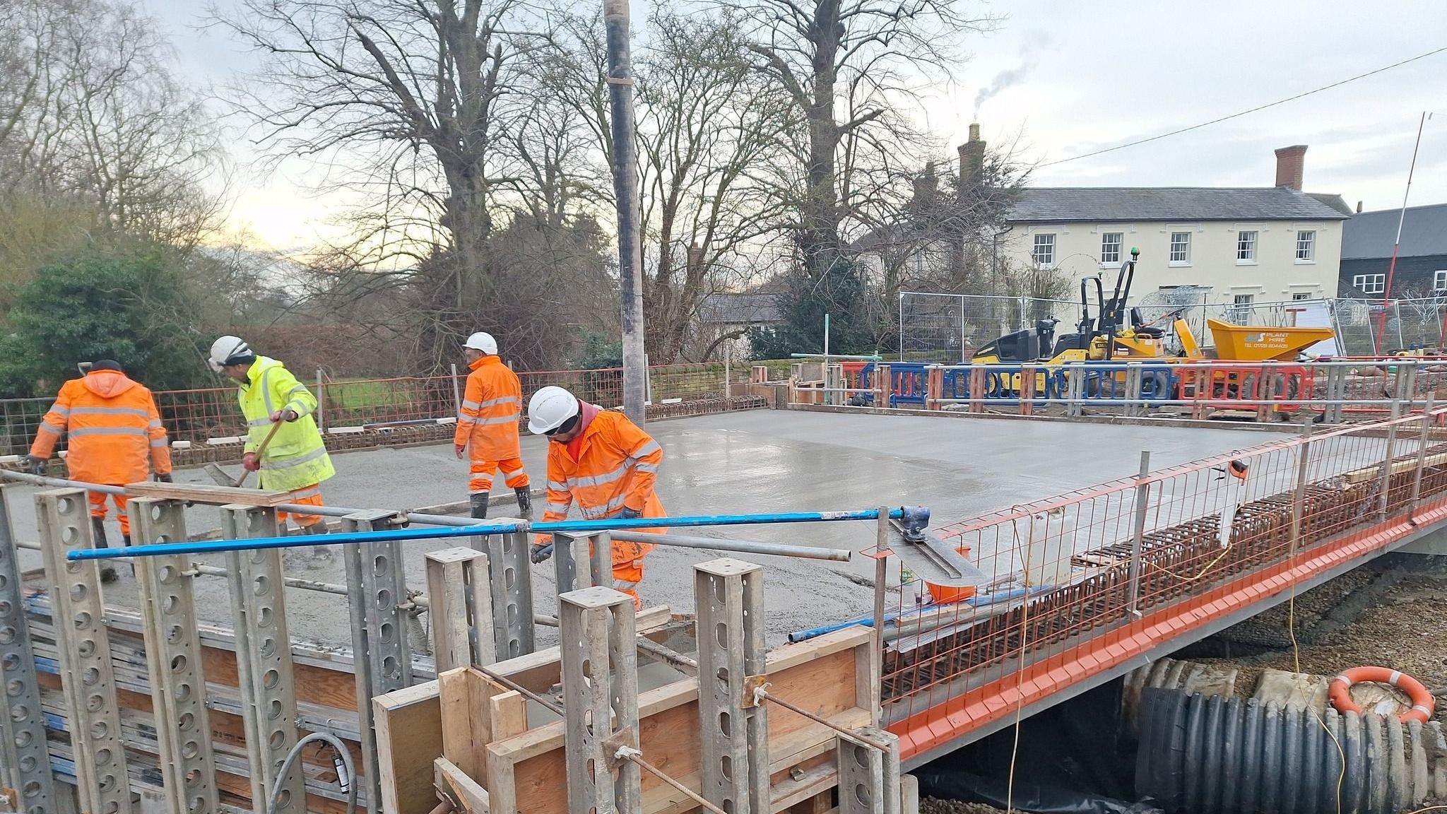 Highways workers wearing orange or yellow hi-vis coats and trousers and  white hard hats work on a new bridge. The area has been cordoned off and machinery can be seen in the distance.