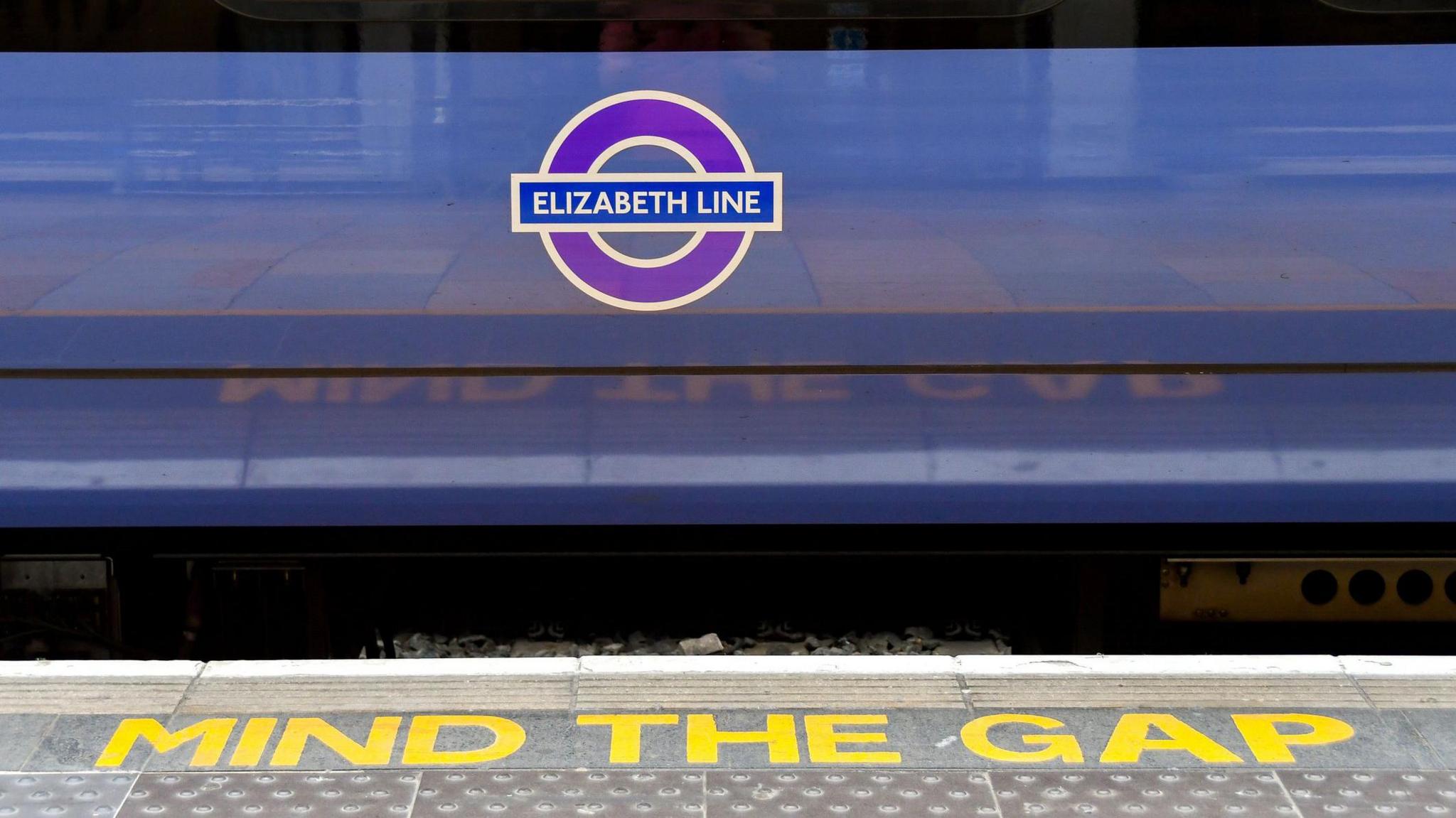 A close-up of a train with a purple "Elizabeth Line" roundel on its side. The platform edge has "MIND THE GAP" written in bold yellow letters. 