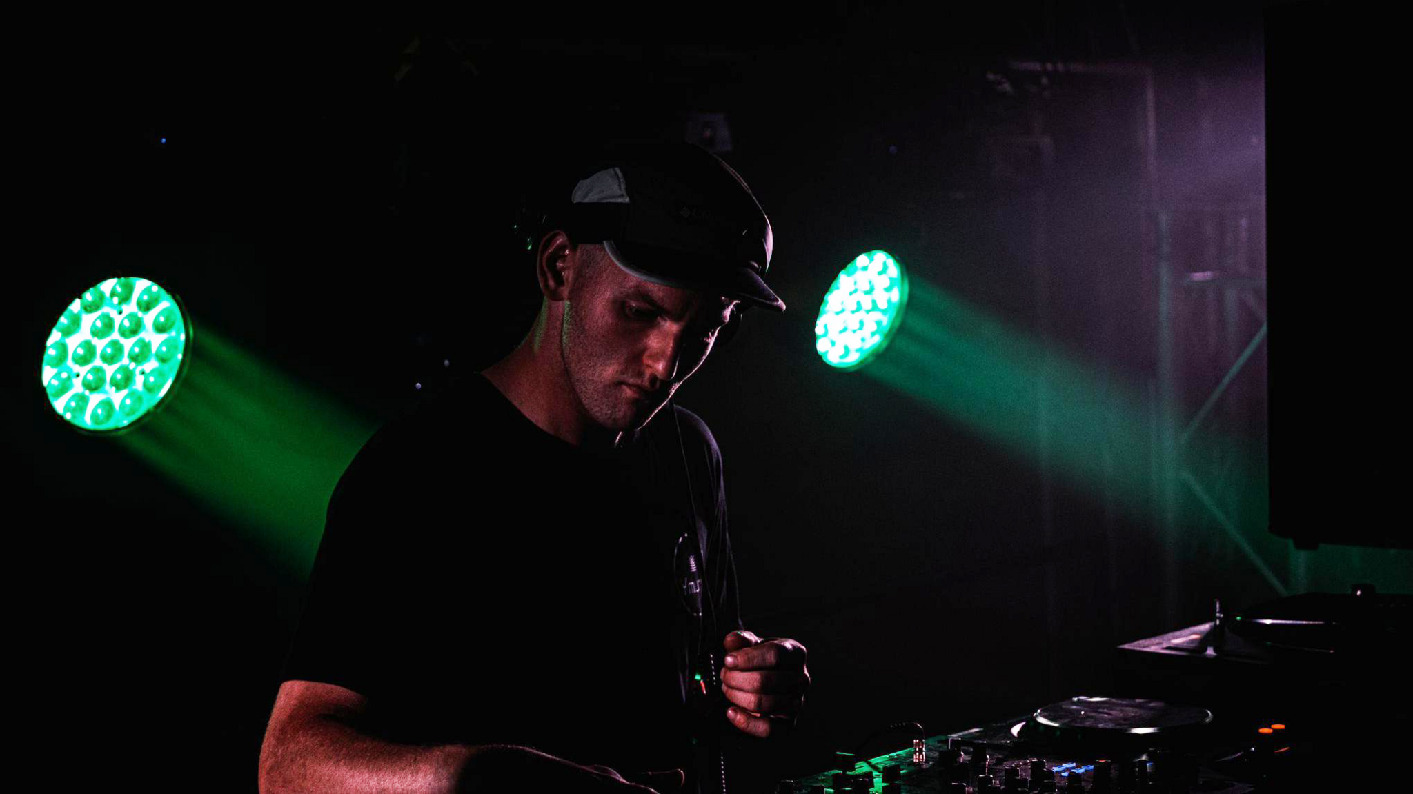 A man wearing a baseball hat, standing in front of DJ decks. The room is dark, and there are two green lights shining down on him.