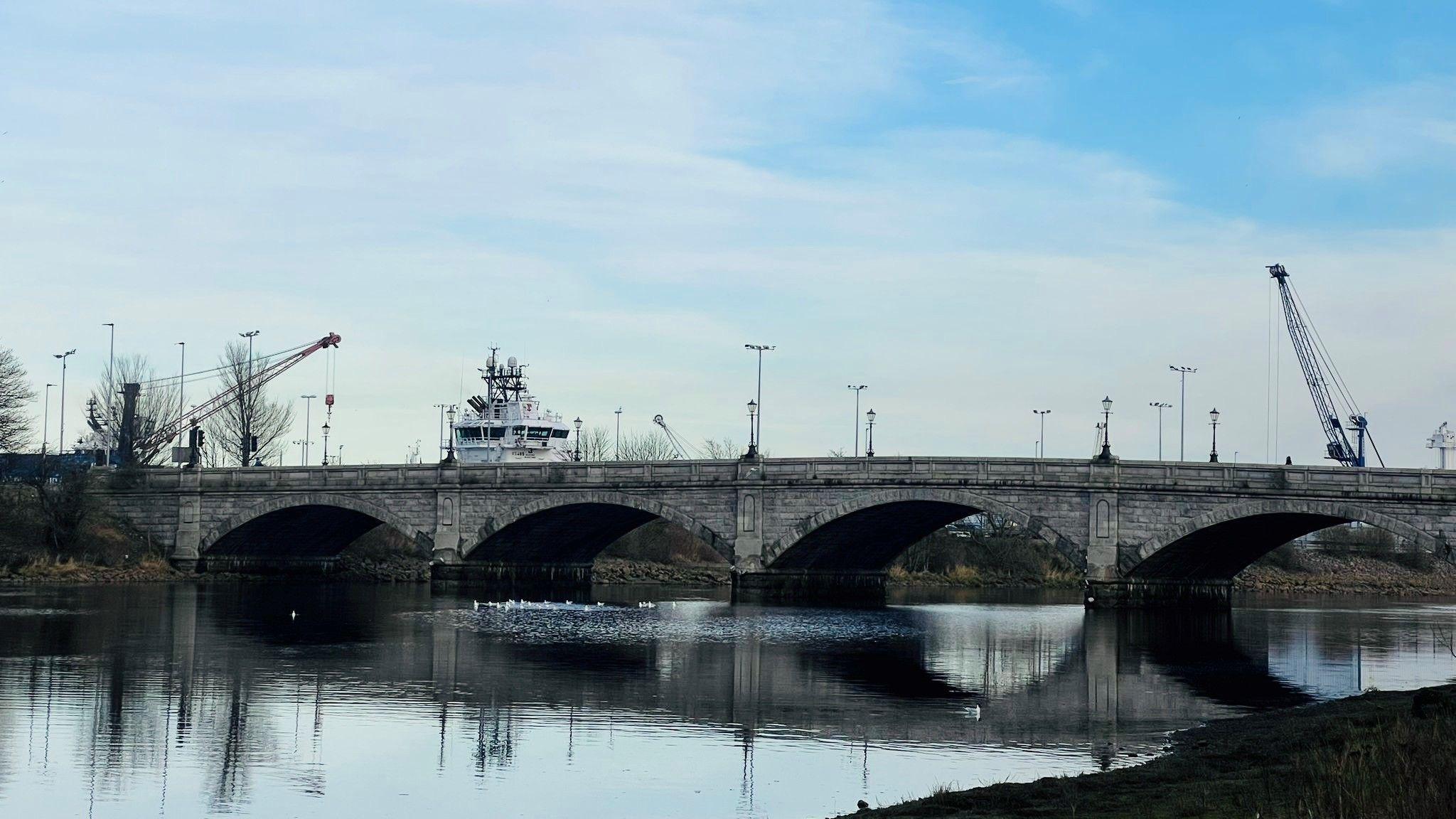 Bridge with oval arches across a river with birds on the water and a ship in the background, against a cloudy blue sky.