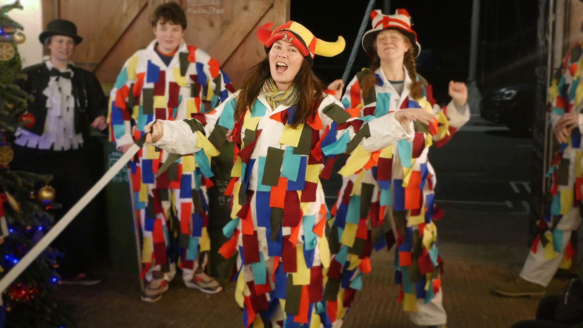 One boy and two young women in white costumes covered in multi-coloured ribbons, standing in a enclosed yard with wooden doors. The Woman at the front is wearing a horned hat, which the other woman is wearing a hat with the English flag on it.