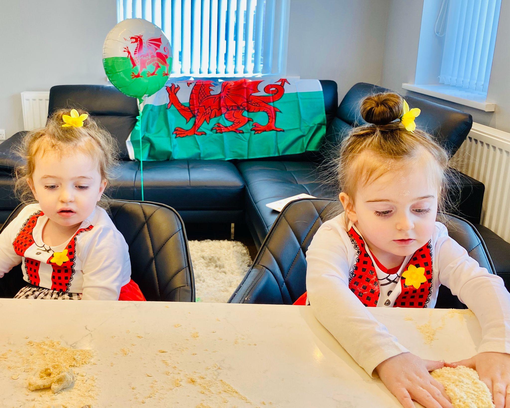 Twin eight-year-old girls wearing Welsh lady costumes of white and red tops and red skirts, with daffodils in their tied up hair, sit at a kitchen island looking down. A Wales flag is on display on a sofa in the background, and next to it is a helium balloon which also has a Welsh flag design on it.