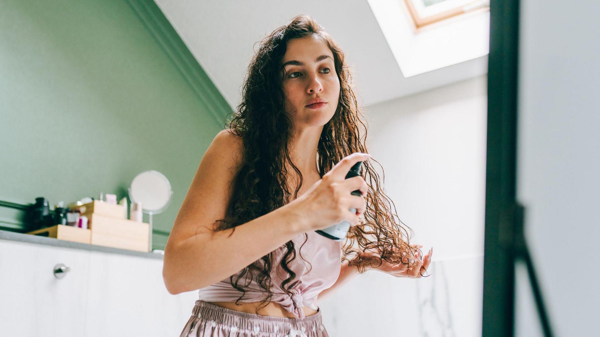 A woman with long brown hair spraying a hair product onto her hair.