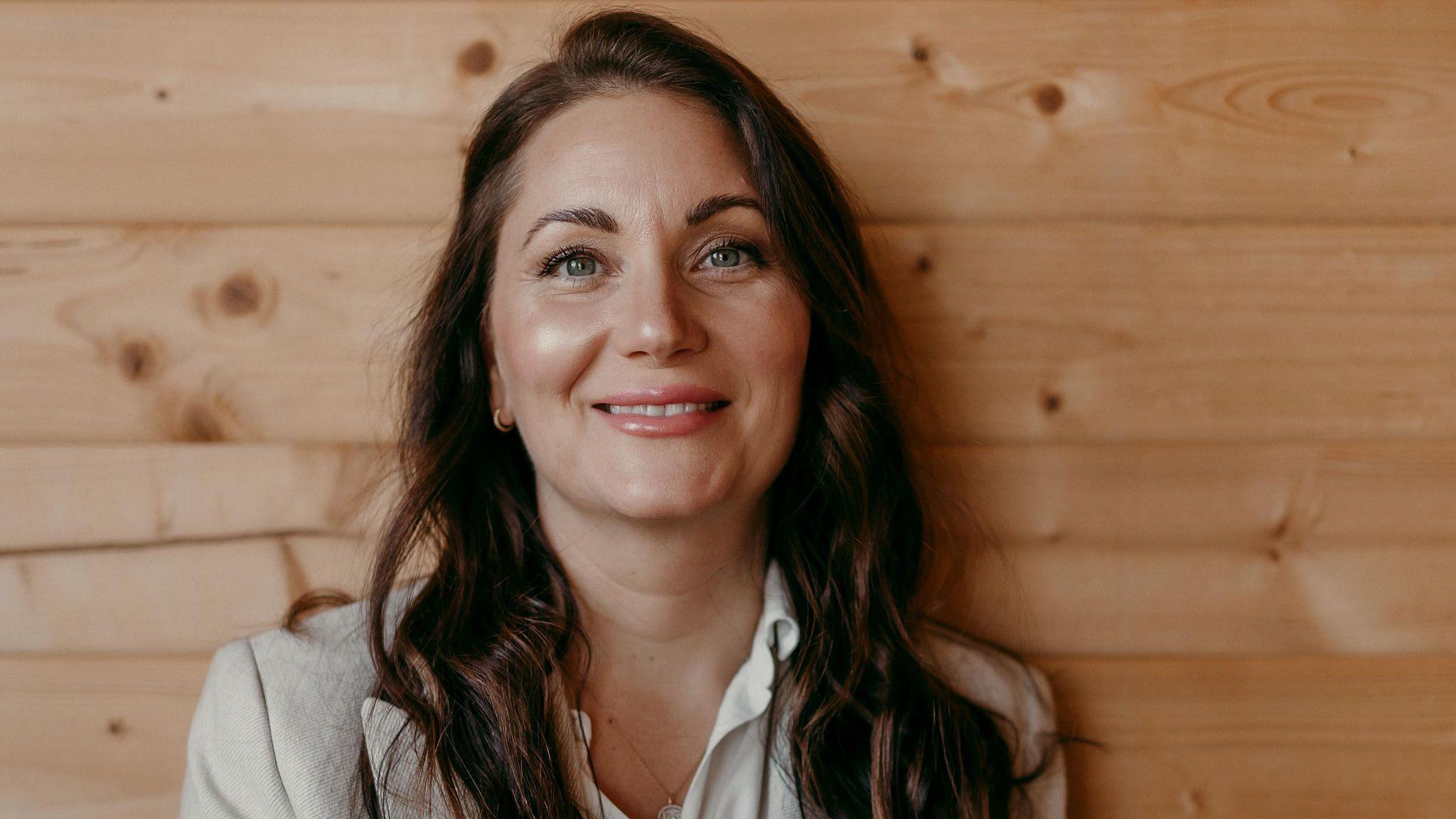 Natalie Boll has long brown hair and is wearing a grey blazer.