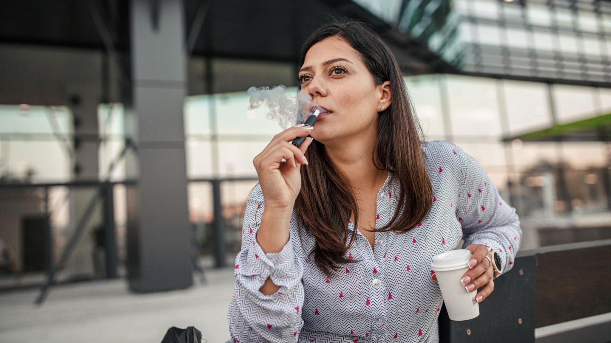 Young woman with brown hair vaping. She has a coffee cup in one hand and is sitting outside a glass-fronted building. 