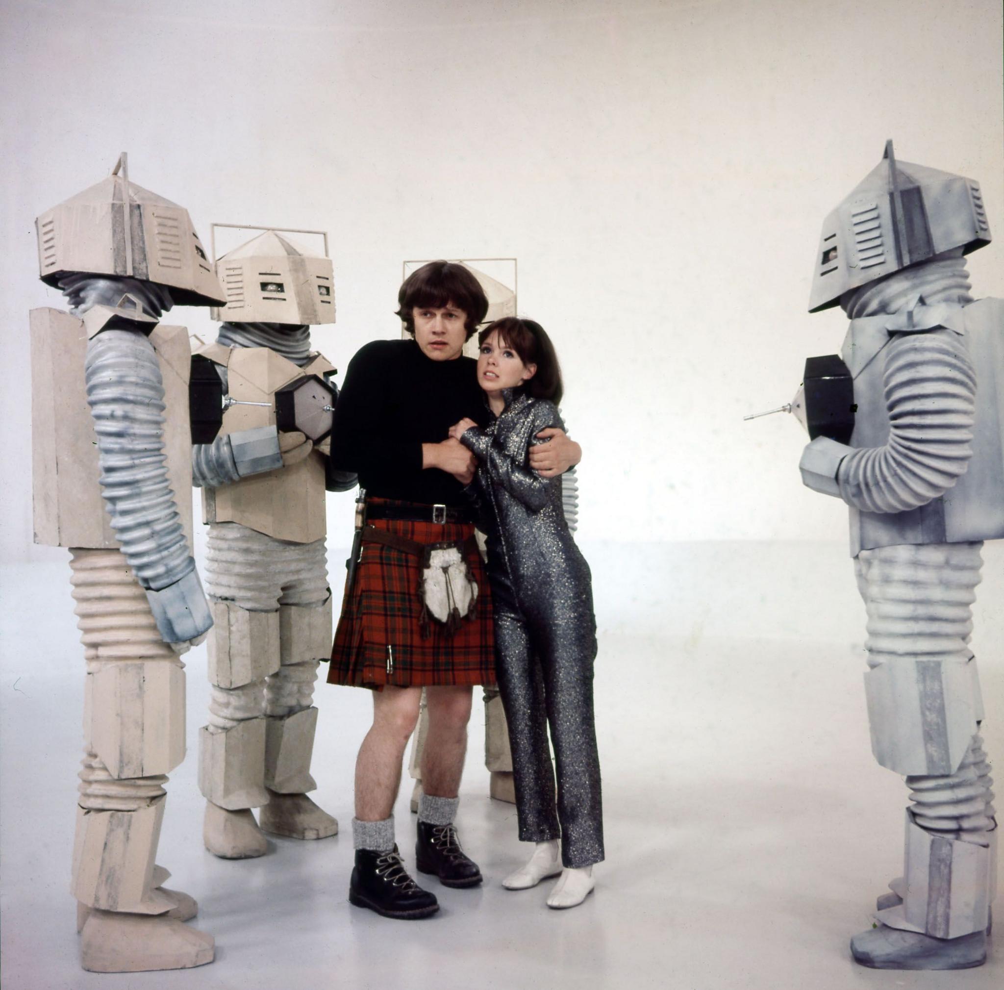 Colour photo of Frazer Hines and Wendy Padbury huddling together, surrounded by large white robots