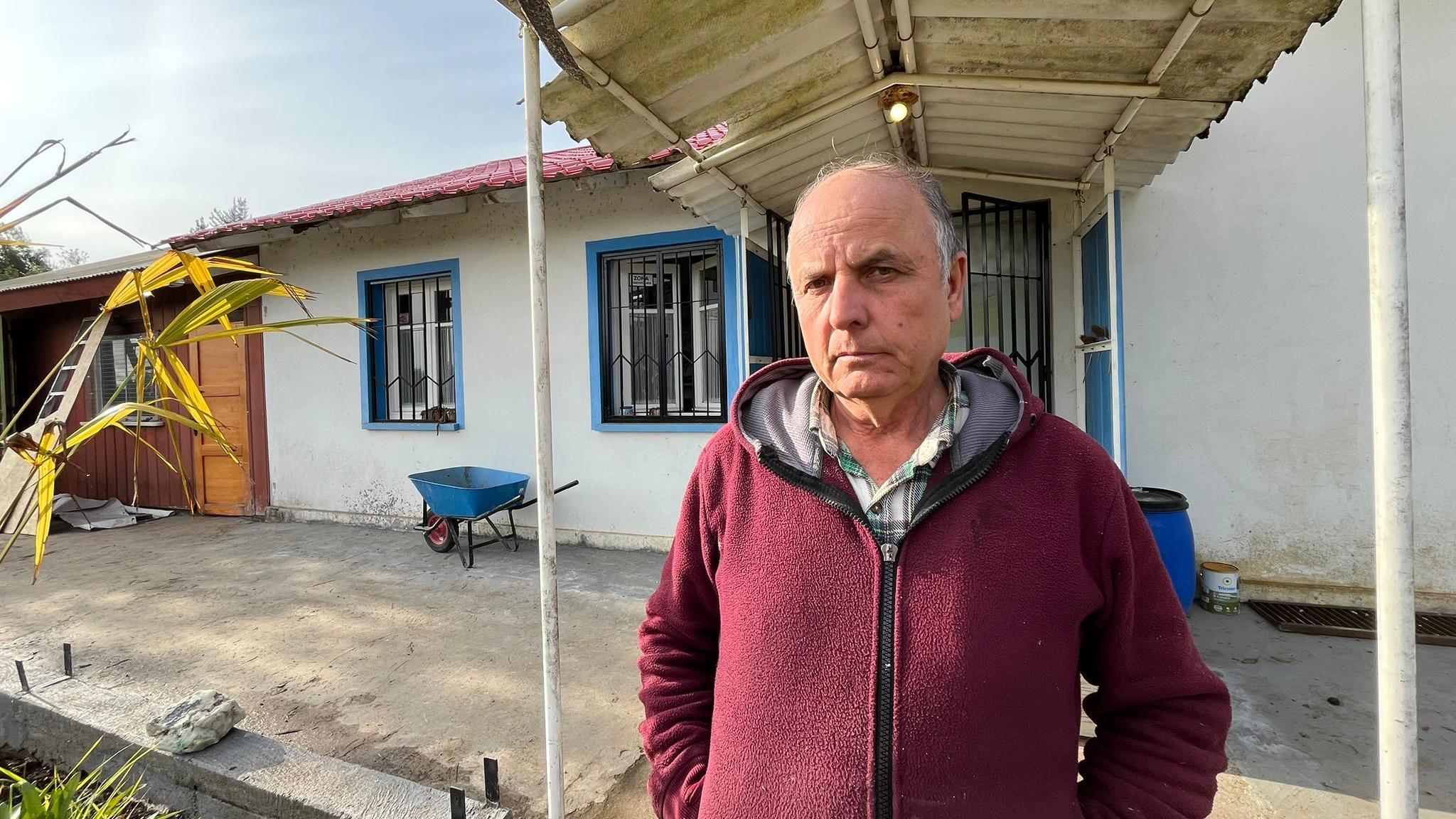 Georg Klaube, in a red hoody, stands in front of a house in Villa Baviera. A blue wheelbarrow can be seen standing in front of a wall. The house is painted white and has blue accents around the windows.  